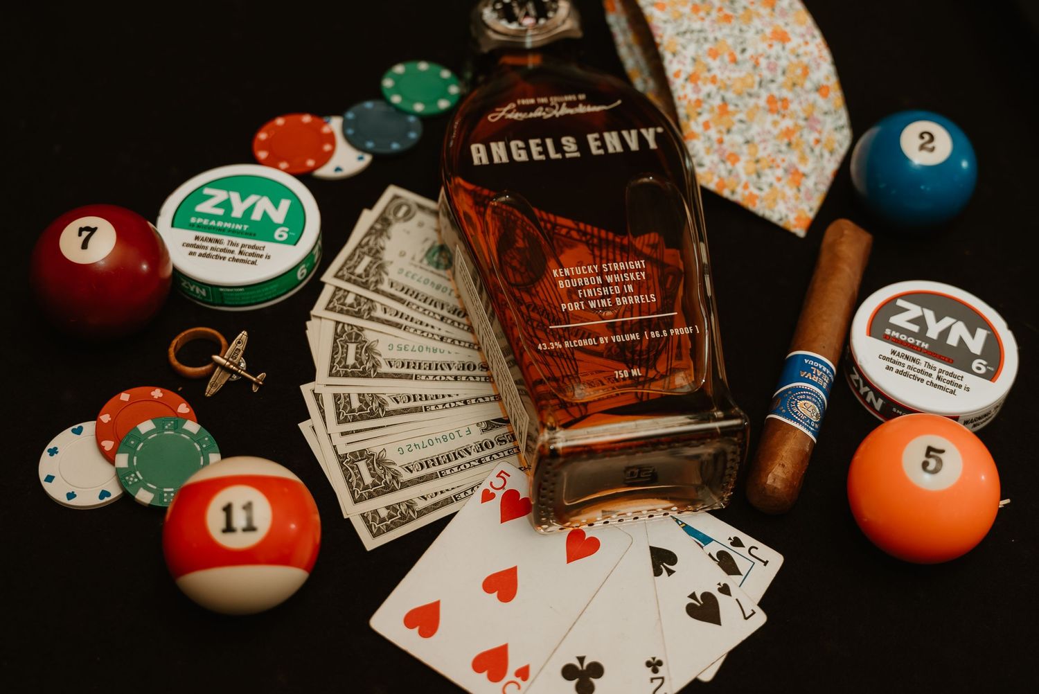 Angels Envy bourbon bottle surrounded by poker chips, playing cards, billiard balls, and cash on dark surface.