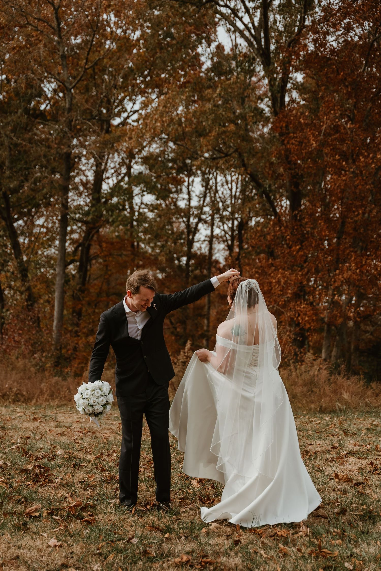 Groom spinning bride in white wedding gown and veil among fall foliage in a forest.