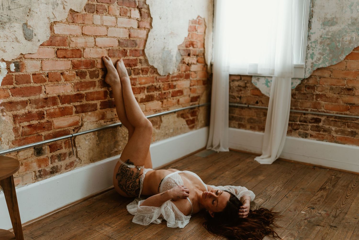 Woman in lingerie and stockings lying on hardwood floor against rustic exposed brick wall in natural light.