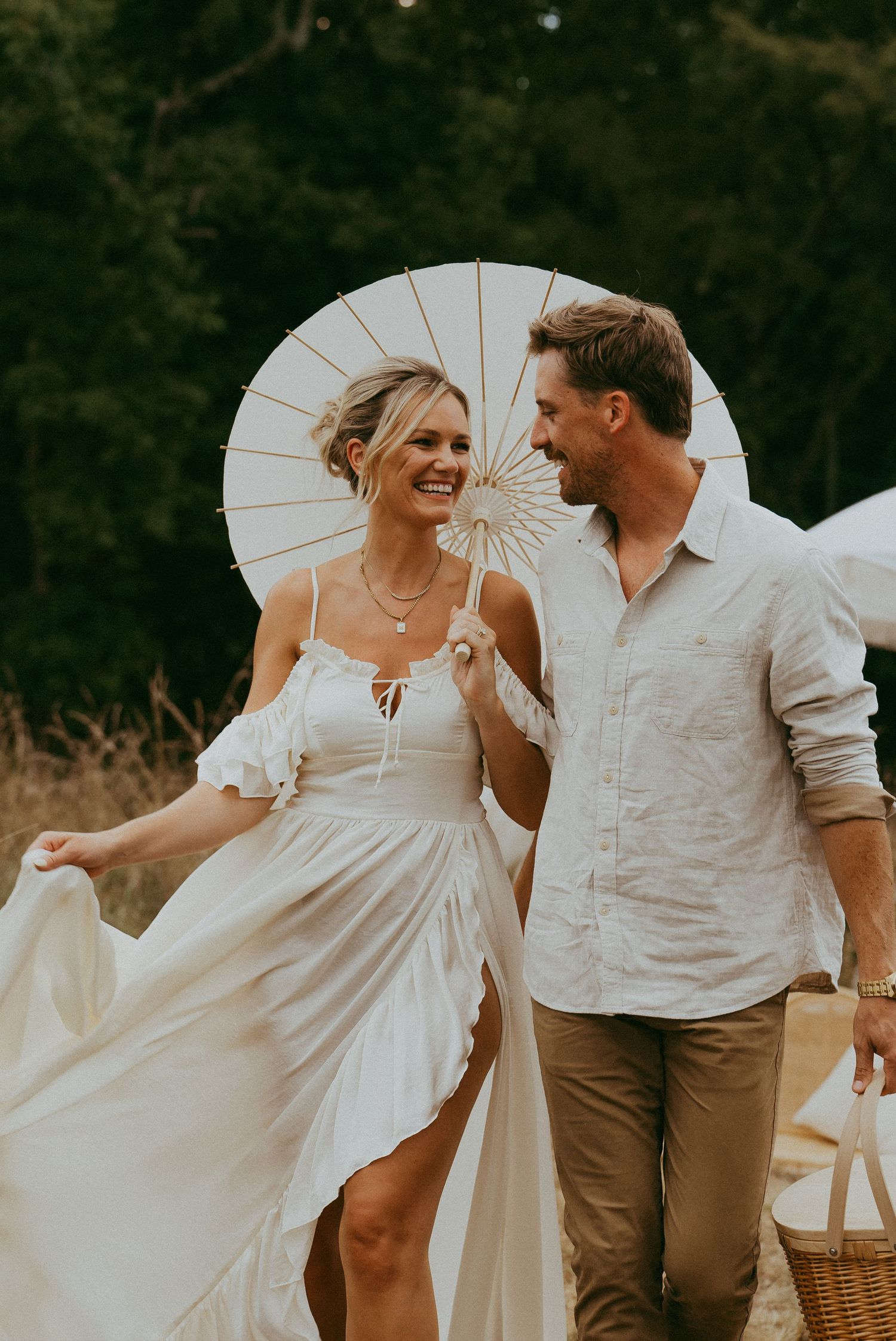 Happy couple walking together under a white parasol in a sunny field during a romantic photoshoot.