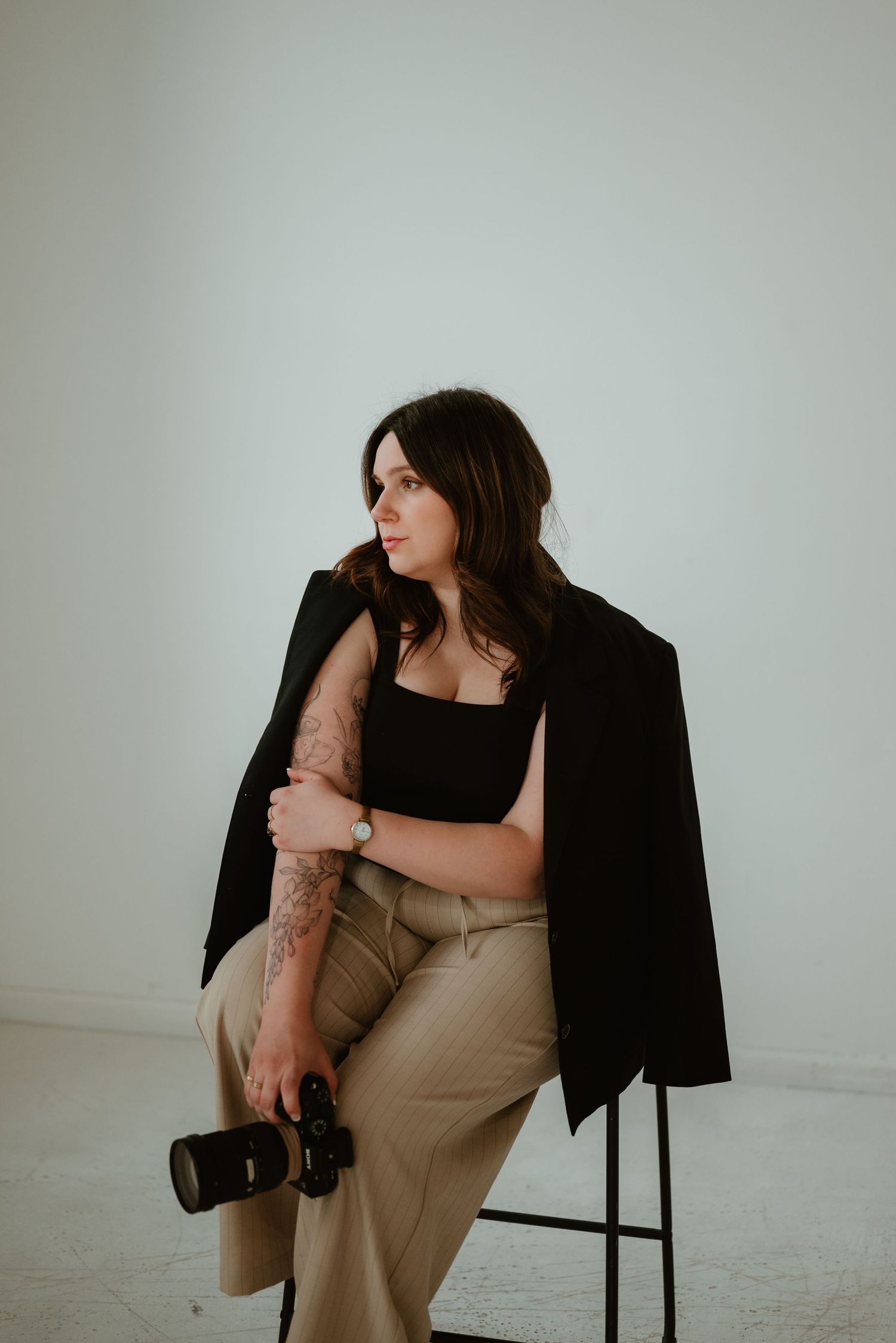 Female photographer sitting on stool holding Sony camera wearing black blazer and tan trousers.