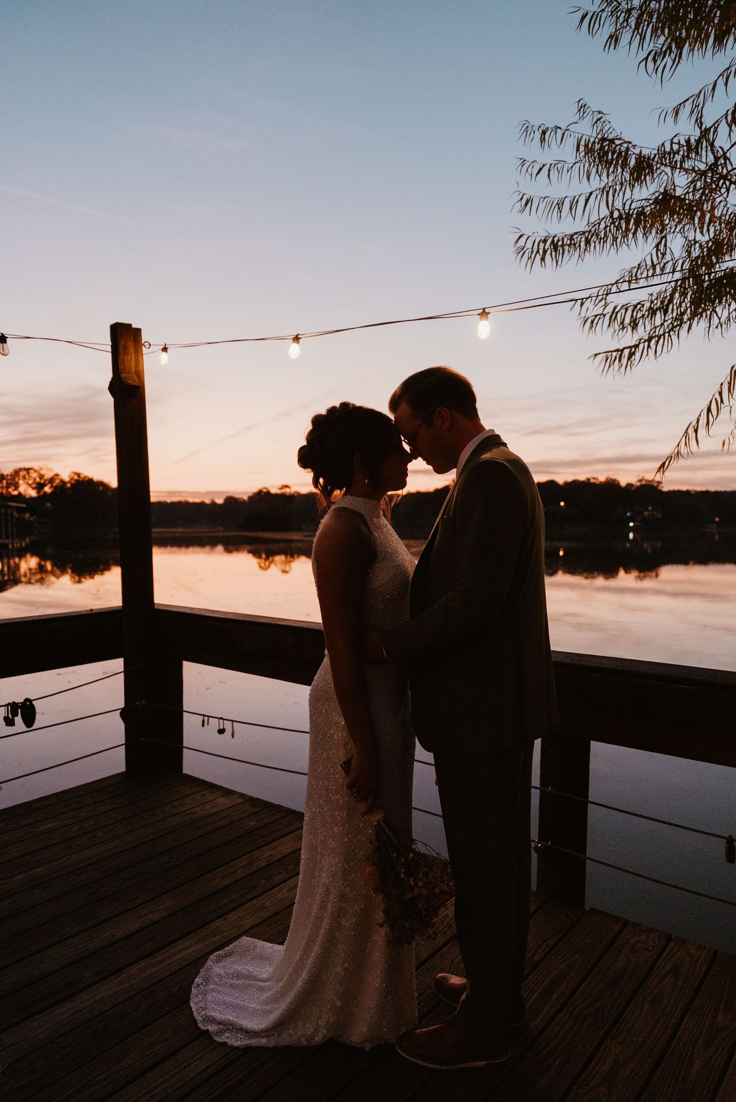 Bride and groom sharing intimate moment on wooden dock at sunset with string lights over reflective lake.