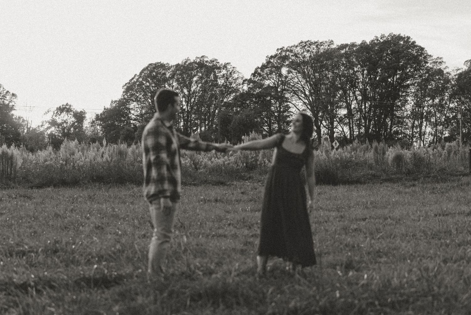 Black and white photo of couple holding hands at a distance in an open field with trees behind.
