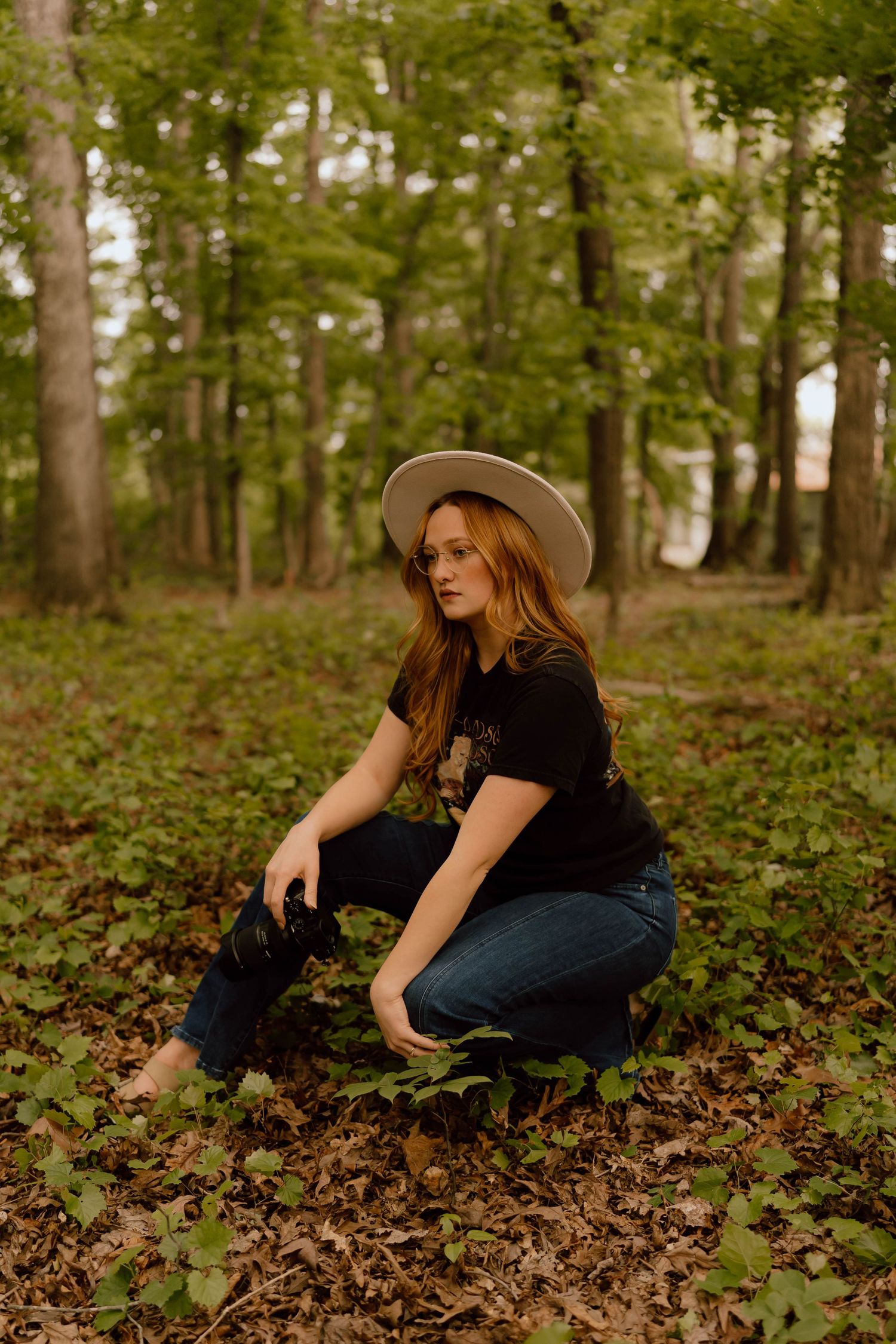 Red-haired woman in black tee and jeans crouching in lush green forest wearing wide-brim hat.
