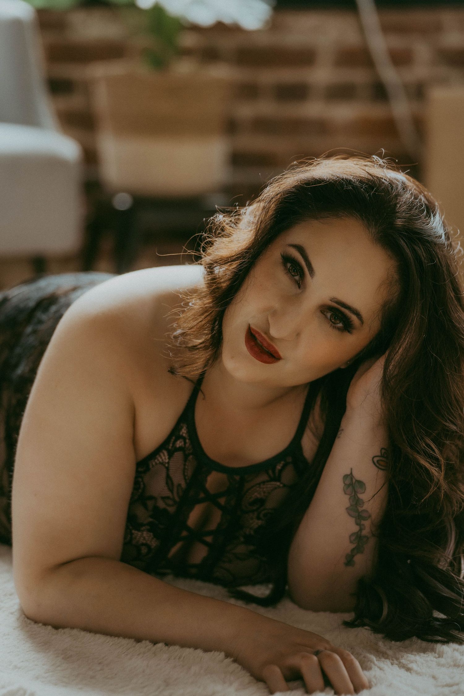 A plus-size brunette woman in black lace bodysuit poses lying down, wearing red lipstick with a tattoo visible.