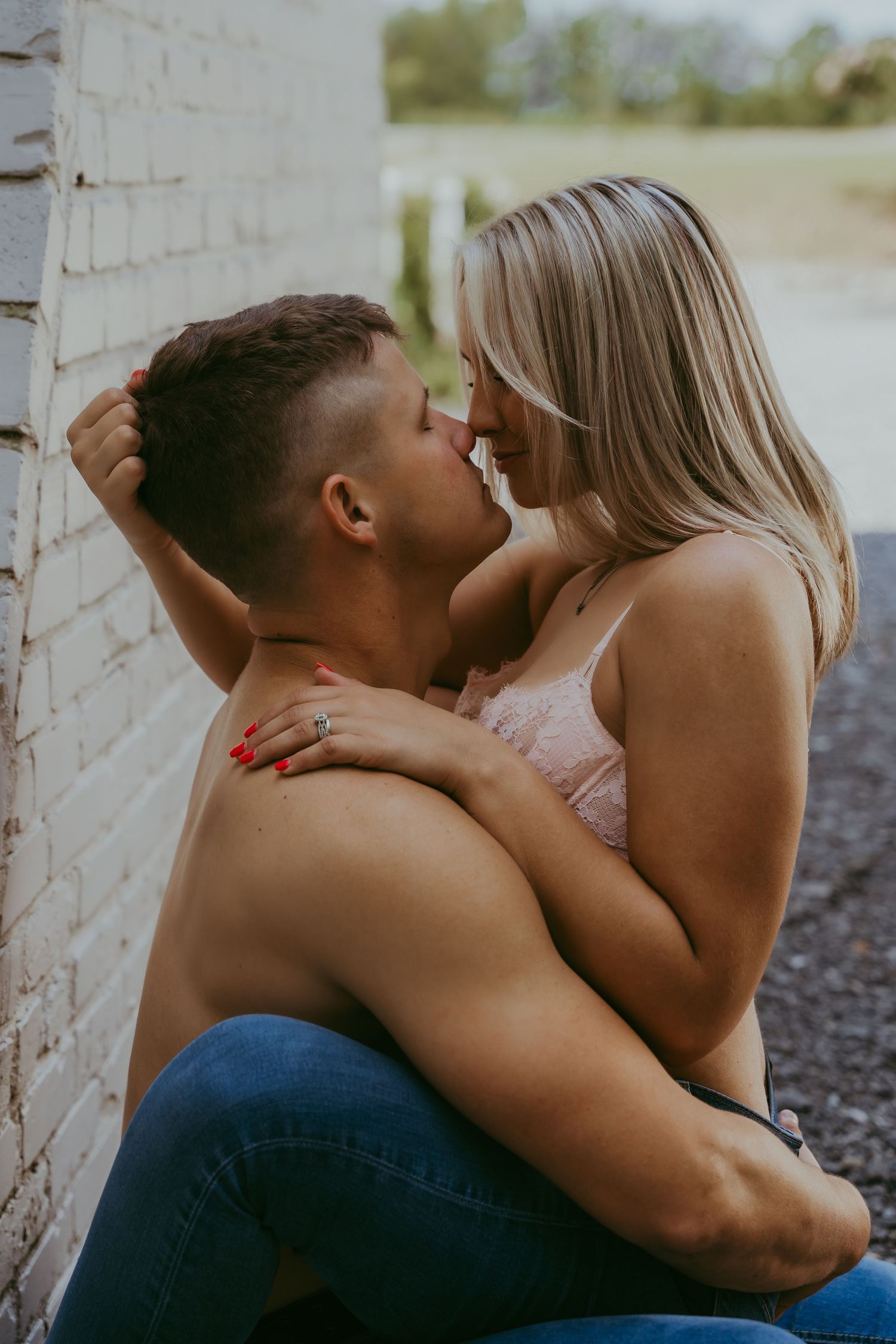A shirtless man and blonde woman share an intimate moment, nearly kissing while embracing against a white brick wall.