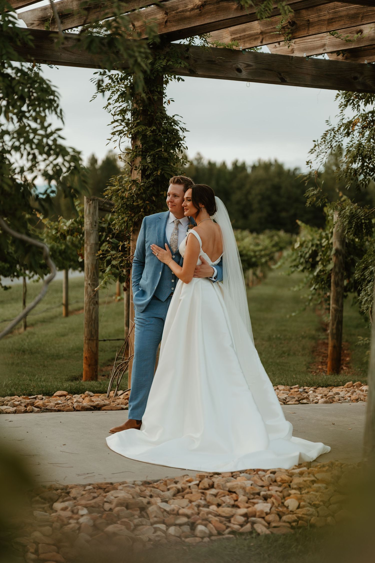 Bride and groom embracing under rustic wooden pergola draped with greenery at vineyard wedding venue.