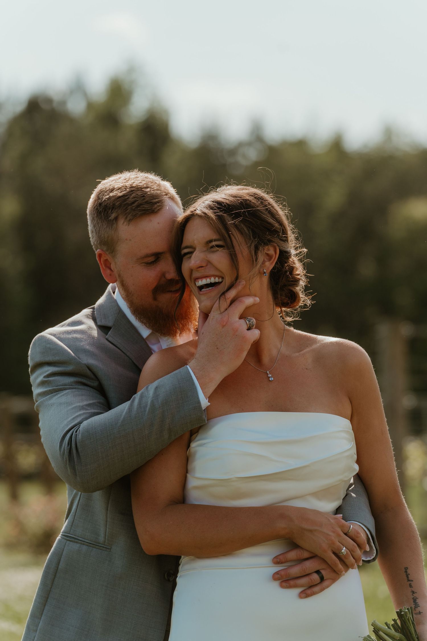 Groom with red beard embracing laughing bride in strapless white gown in golden sunset light.