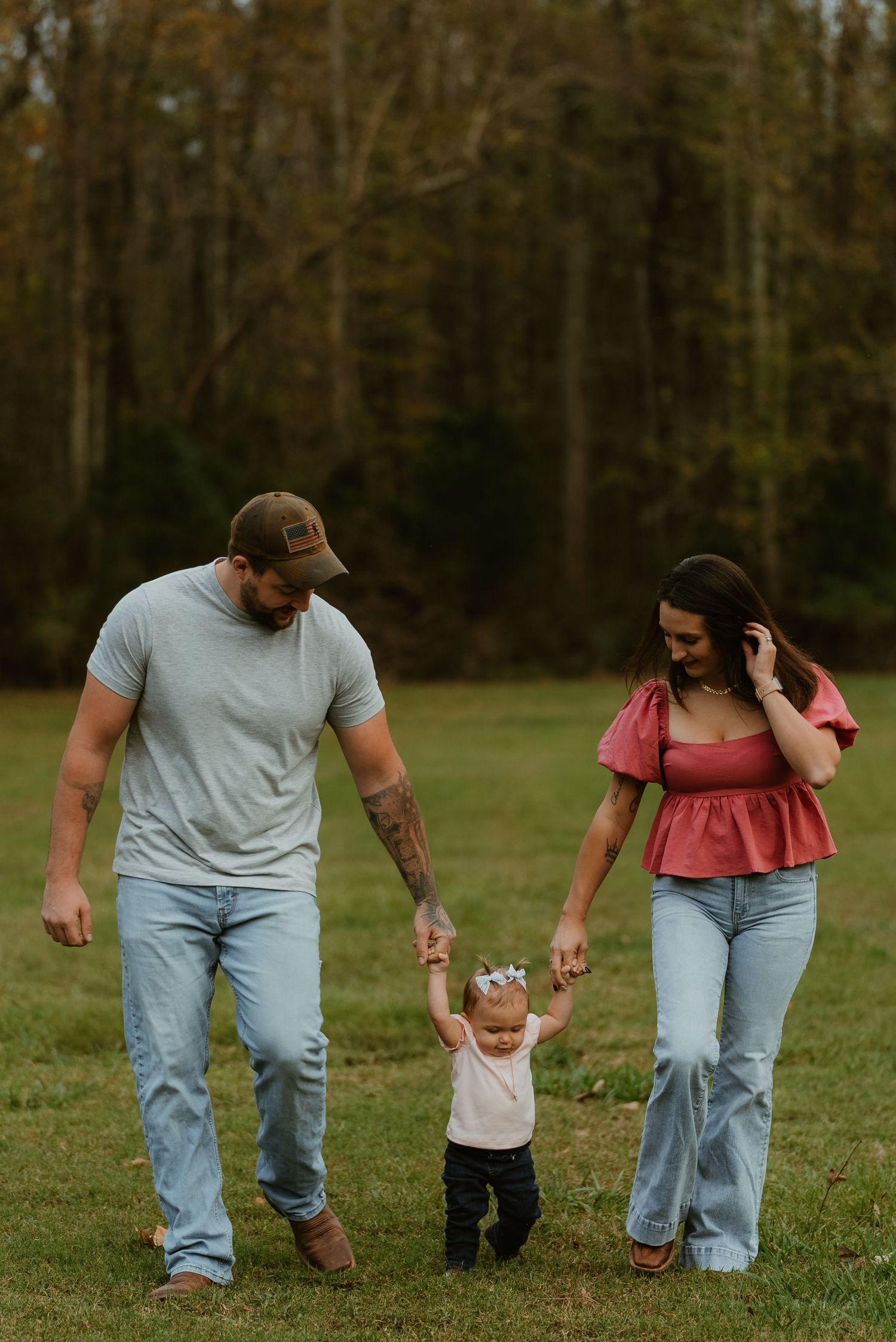 Young parents walk hand-in-hand with their toddler daughter across a green field at dusk.
