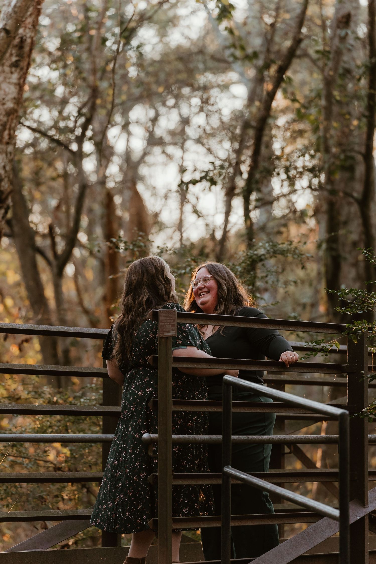 Two women laughing together on a wooden bridge surrounded by autumn foliage in a forest setting.