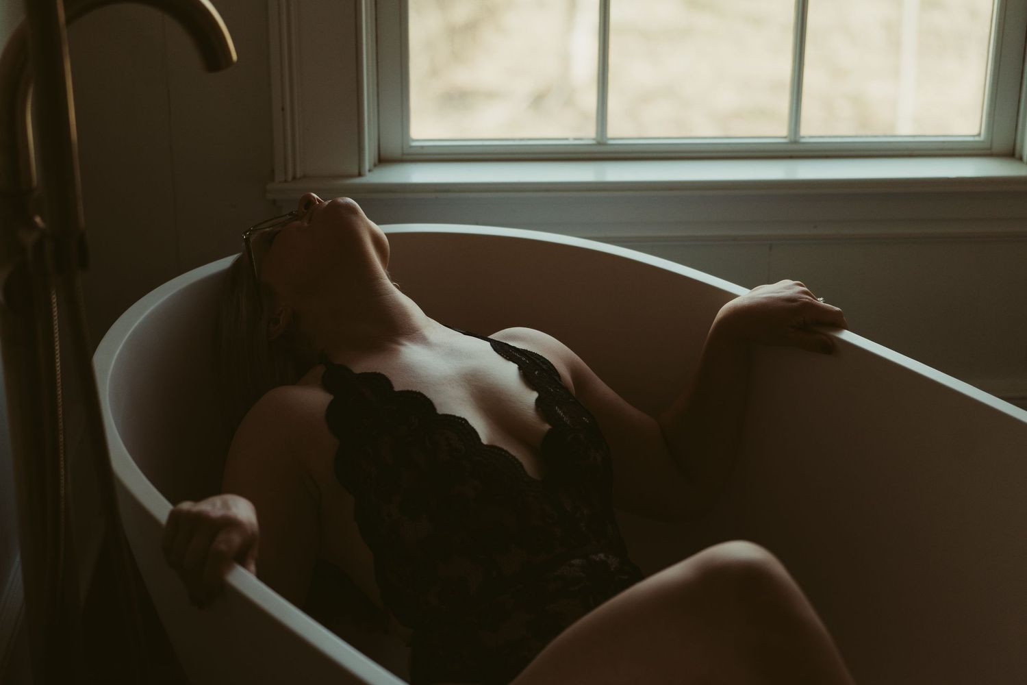 Woman in black lace lingerie reclining in a vintage clawfoot bathtub by a window.