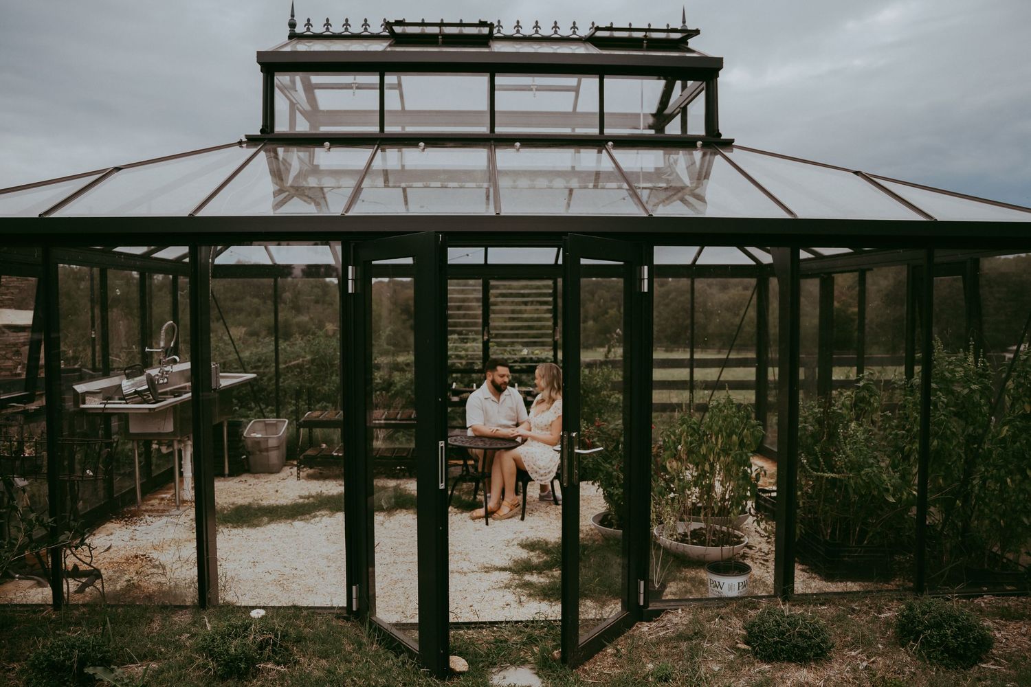Couple sitting together inside a glass greenhouse with cloudy sky backdrop.