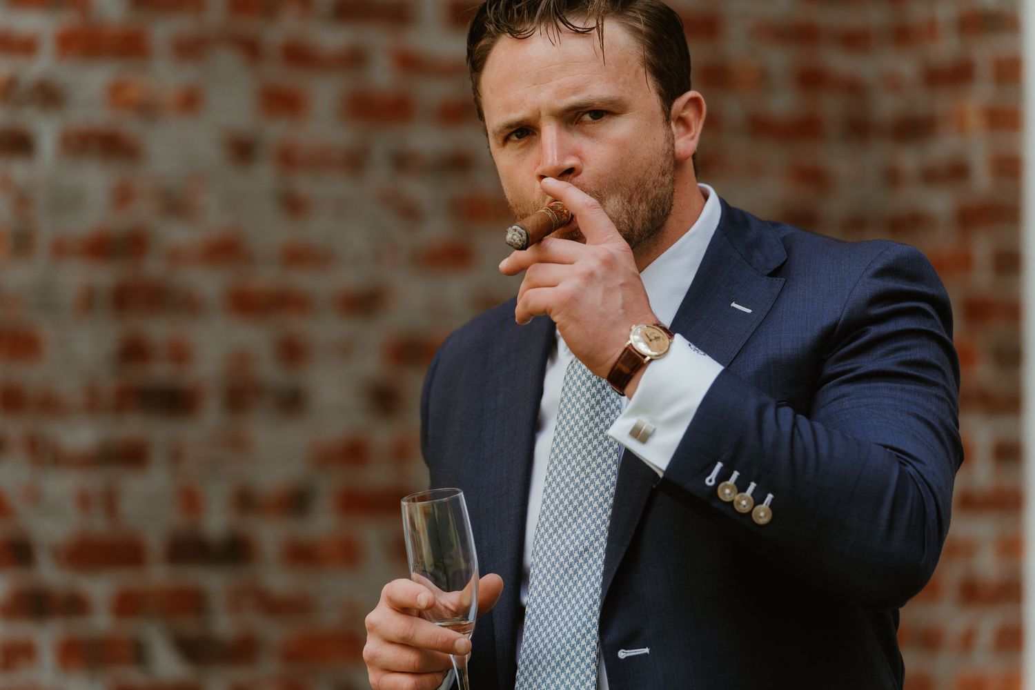 Dapper groomsman in navy suit smokes a cigar and holds champagne glass against a brick wall backdrop.