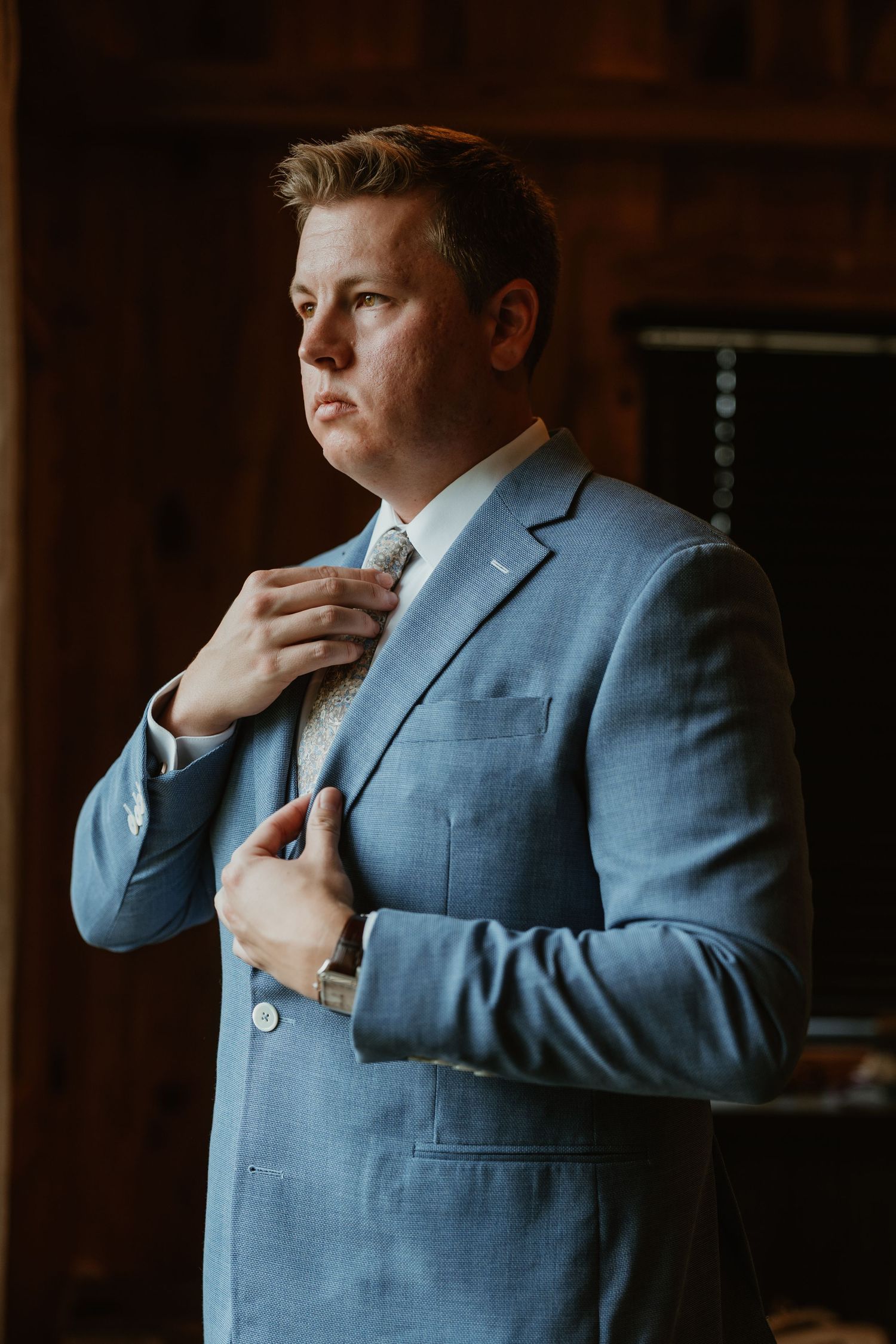 Groom in light blue suit adjusting tie in dimly lit rustic wooden interior before wedding ceremony.