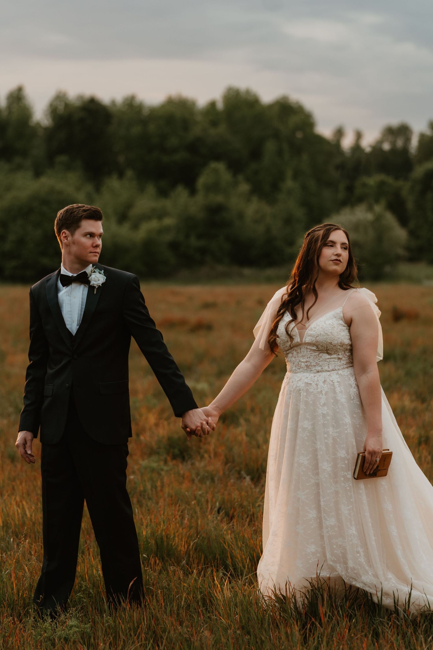 Bride and groom holding hands in golden field at dusk, groom in black tuxedo, bride in sparkle gown.