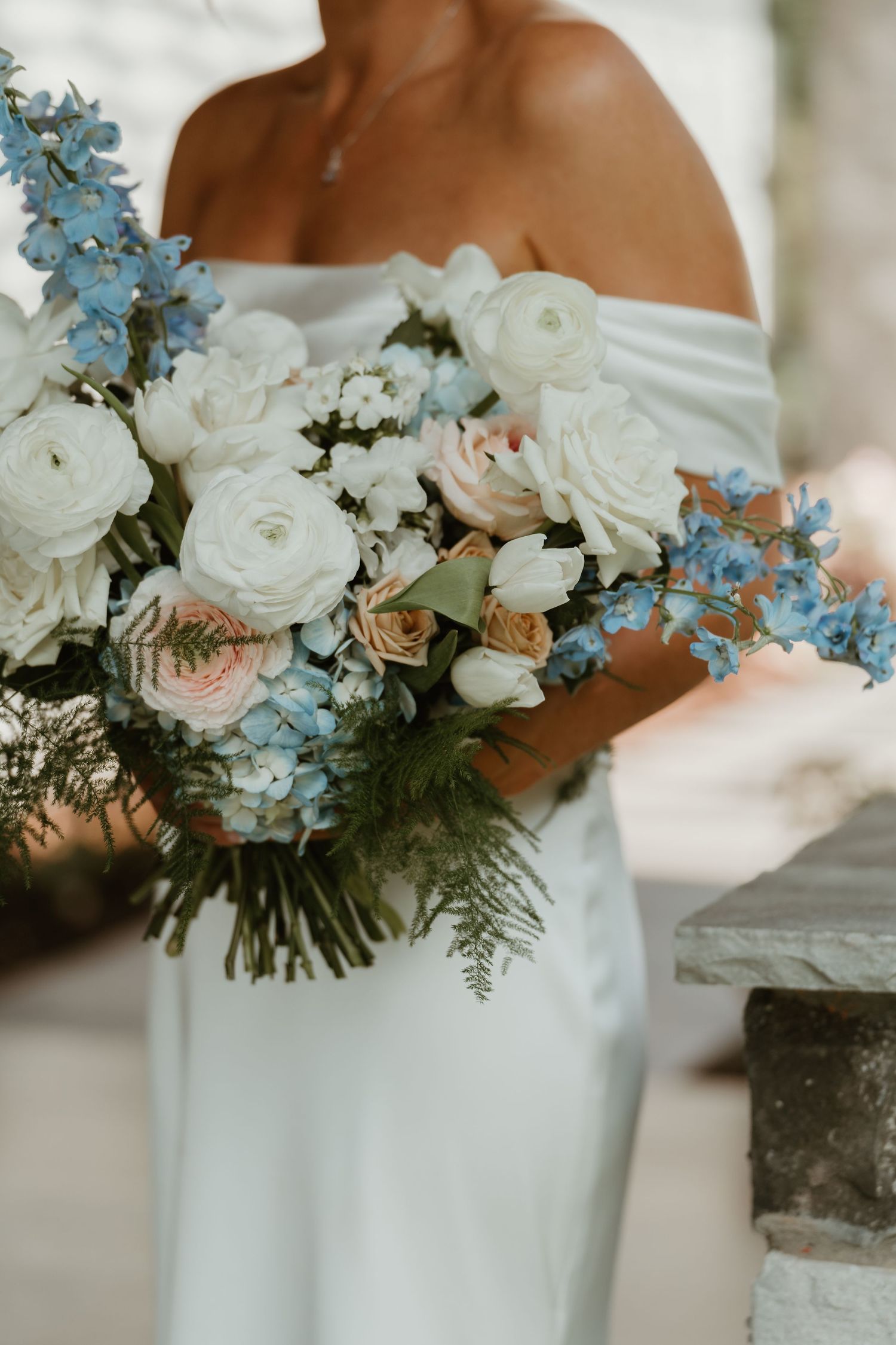 Bride in off-shoulder white gown holding lush bouquet of white ranunculus and blue delphinium flowers.