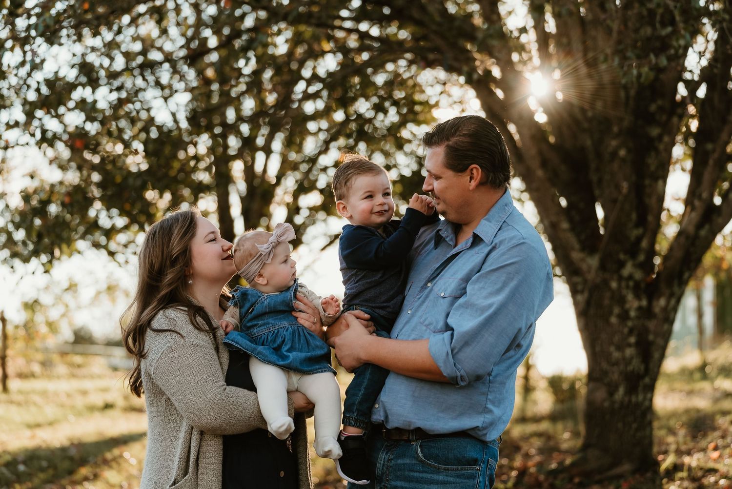 A happy family of four with two young children laughs together in golden sunlight under trees.