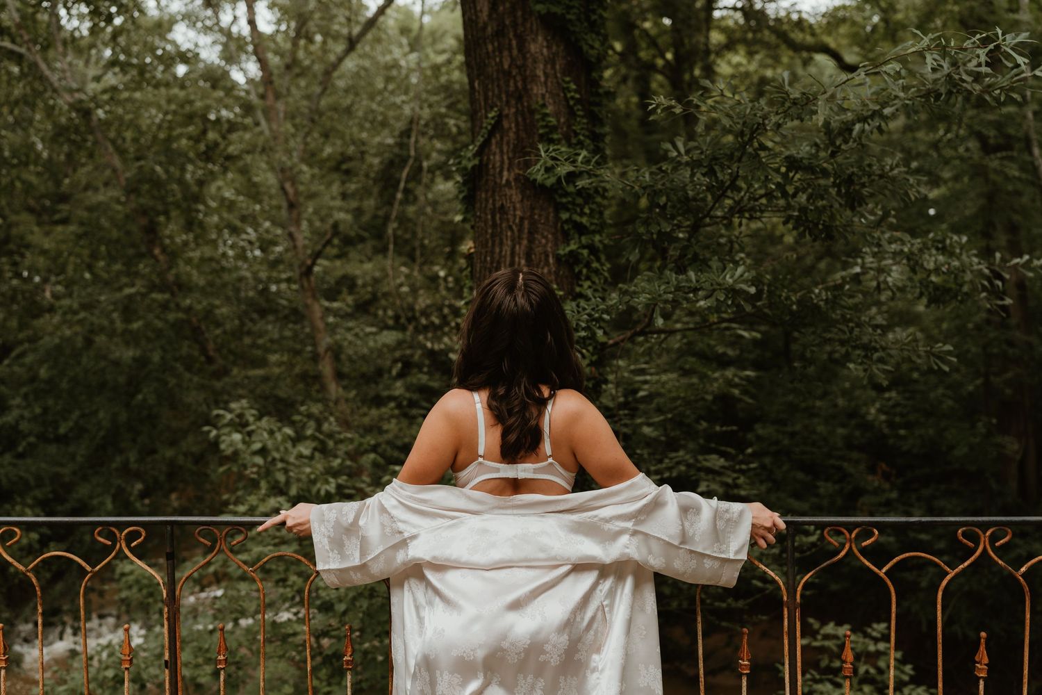 Woman in white robe standing on balcony overlooking lush green forest trees.