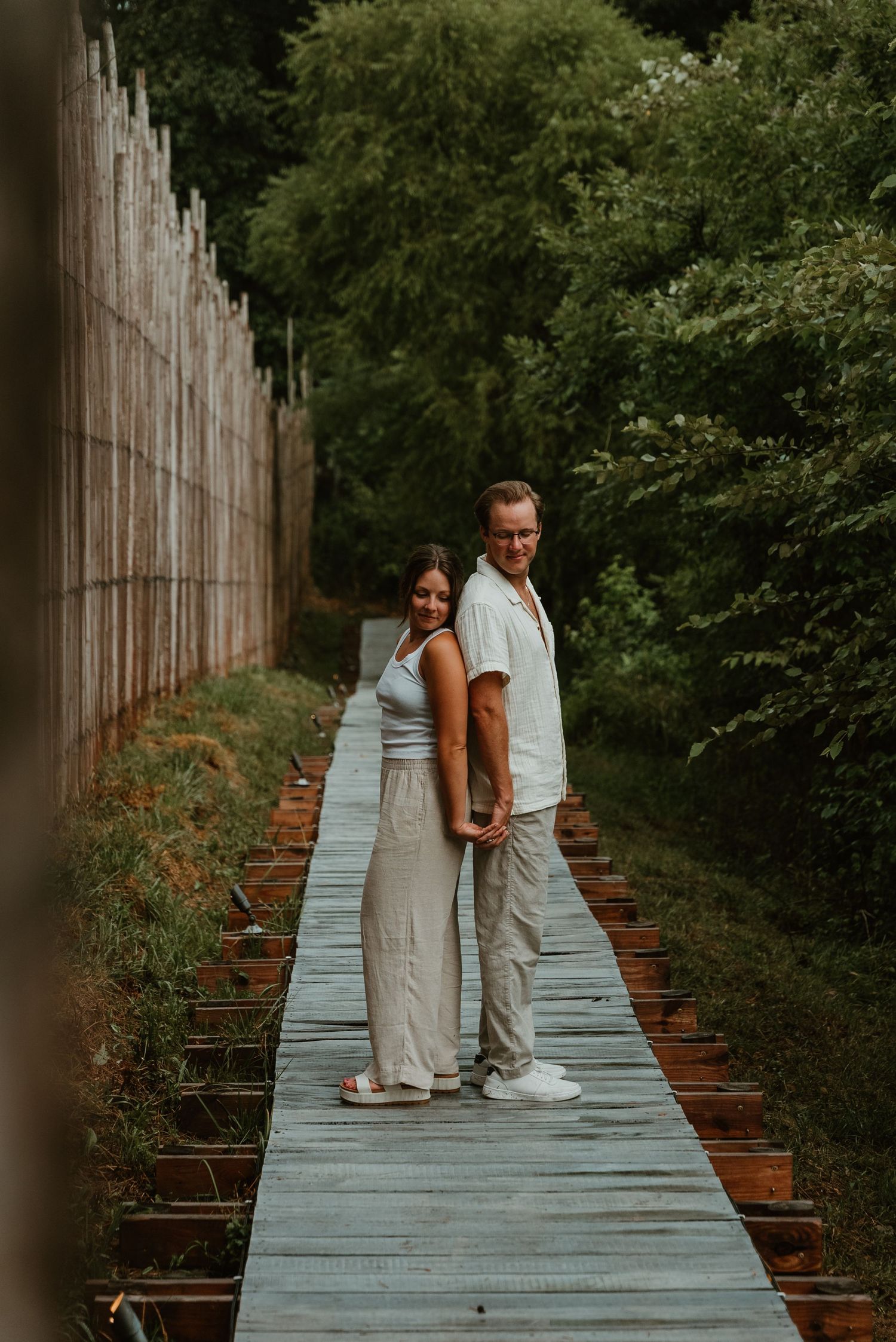 Couple standing on a wooden boardwalk surrounded by lush greenery and bamboo fence during an outdoor engagement session.