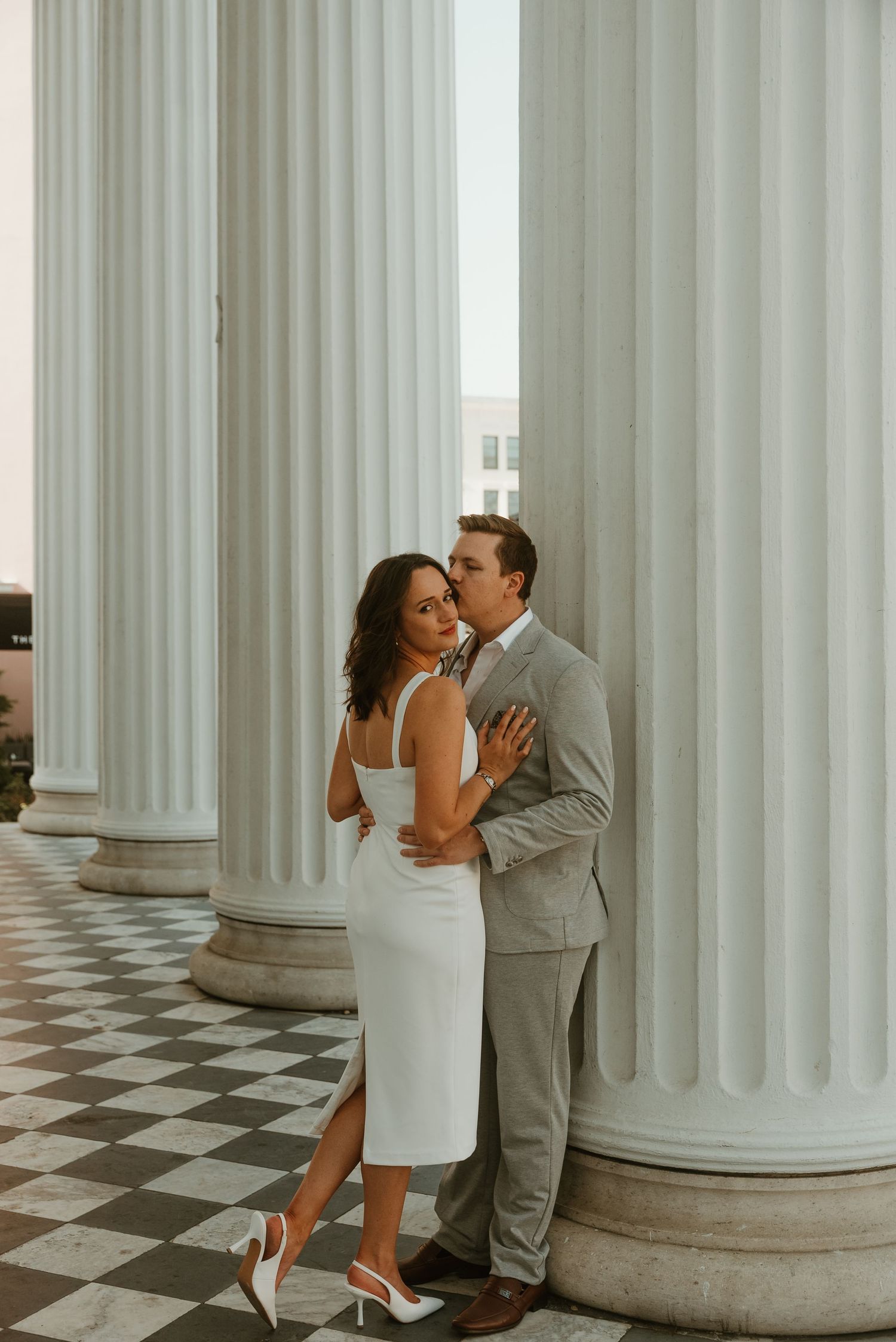 Couple sharing a kiss beside tall white columns in an elegant classical building.
