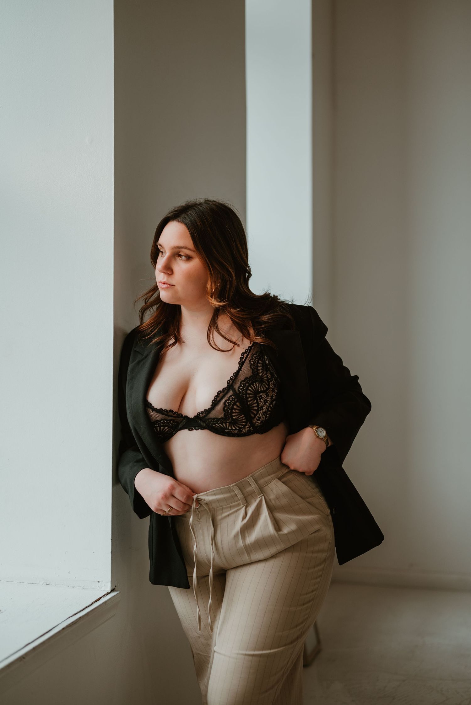 Curvy brunette woman in black lace bralette and blazer with beige trousers posing by bright studio windows.