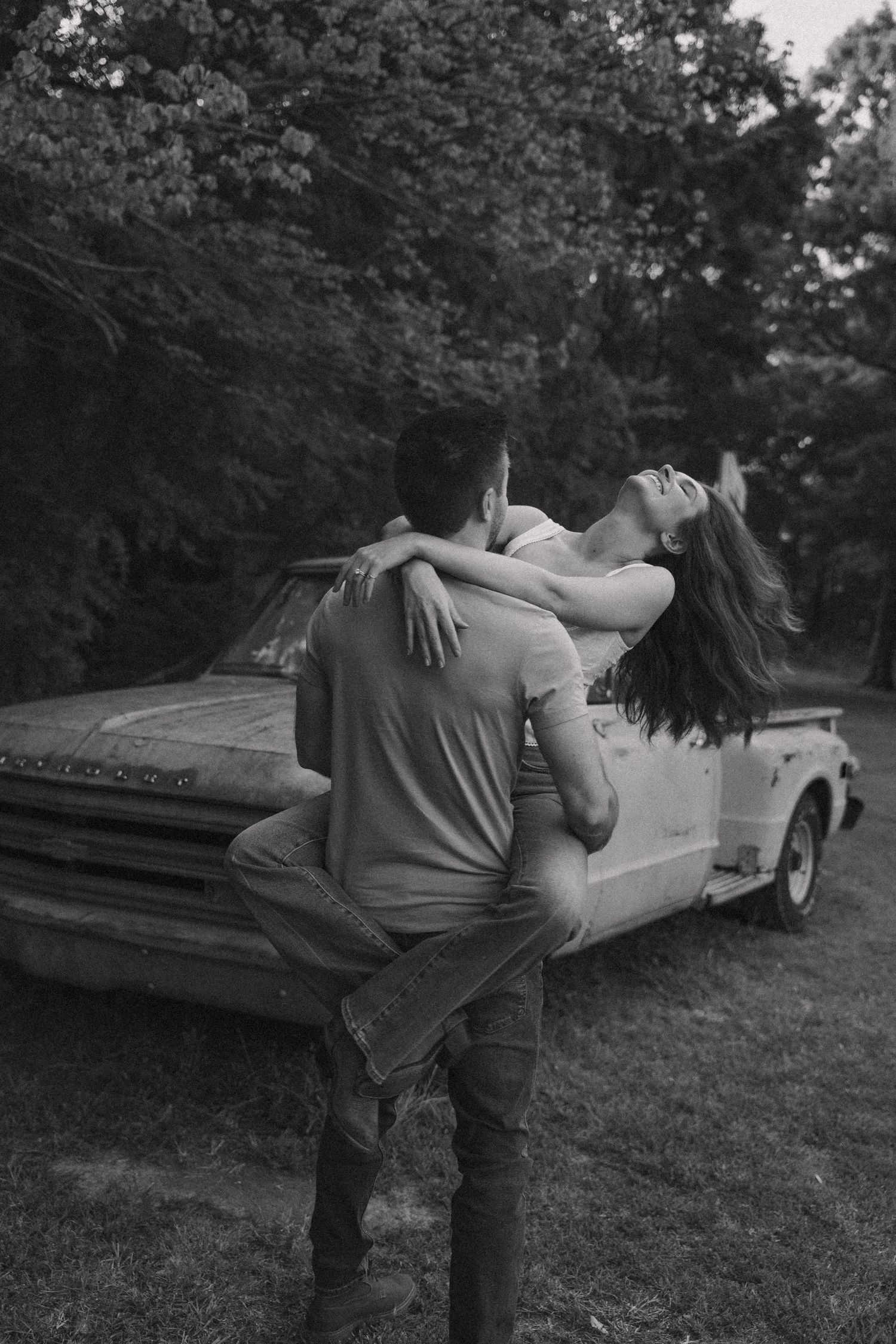 Black and white photo of couple embracing romantically beside a rustic vintage pickup truck.