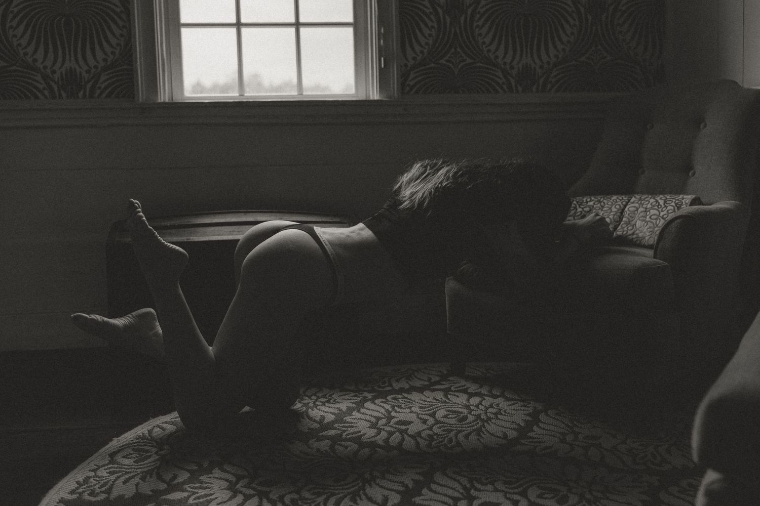Black and white boudoir photo of a woman posing on a rug near a window.