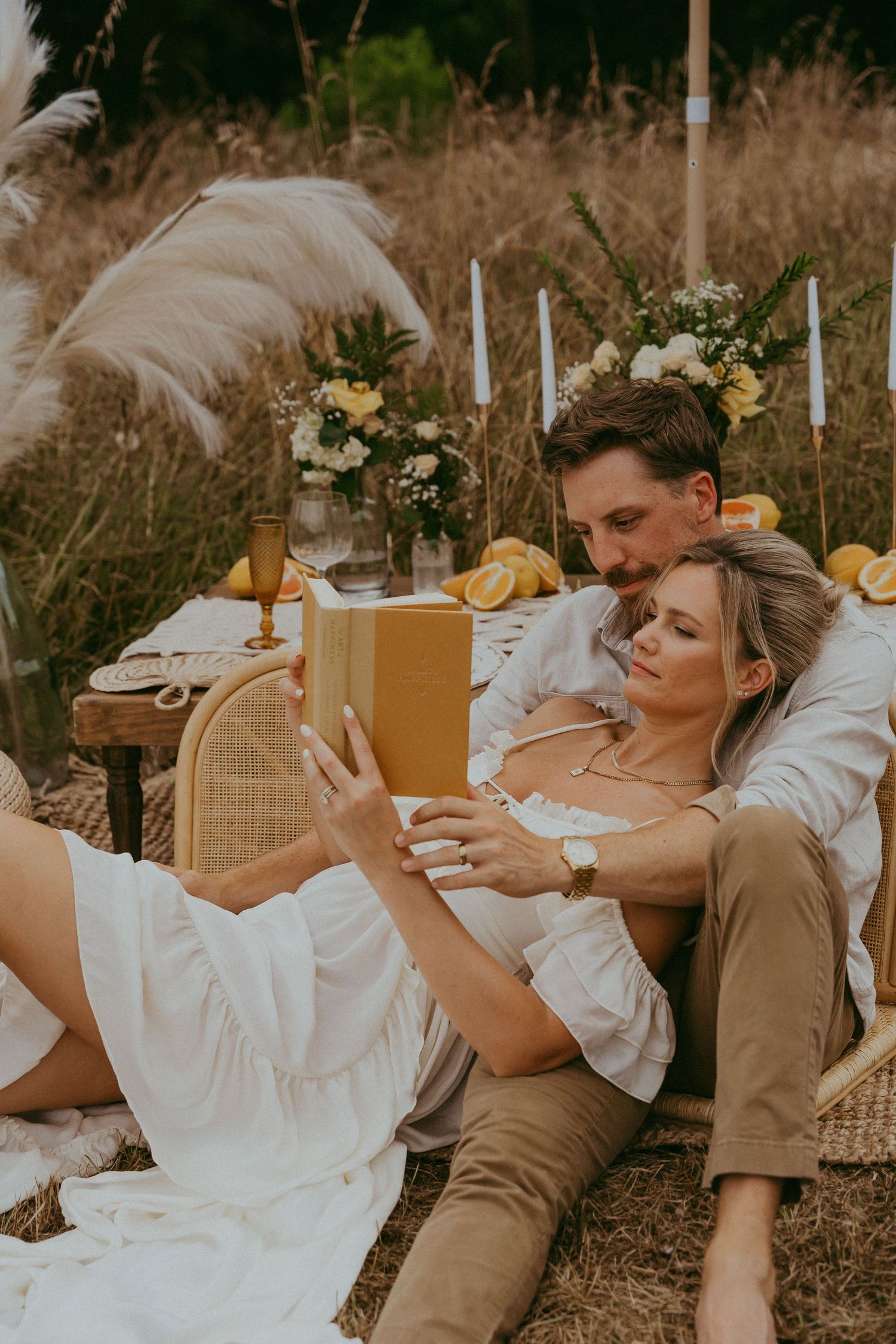 Romantic couple relaxing at a boho picnic in a field, woman reading a yellow book while man kisses her head.