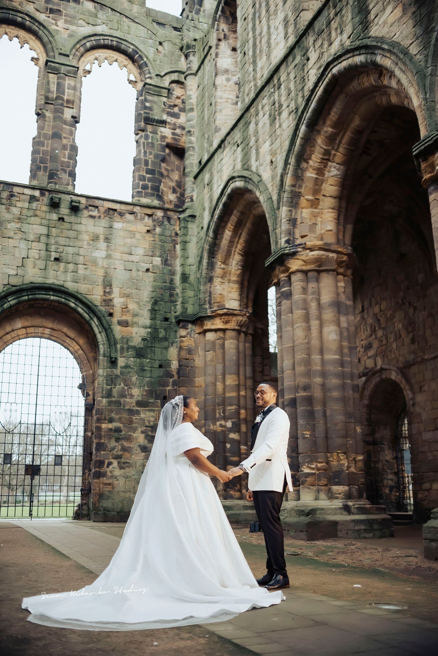 Bride and groom hold hands at historic stone abbey ruins, bride in flowing white gown with veil, groom in white tuxedo jacket.