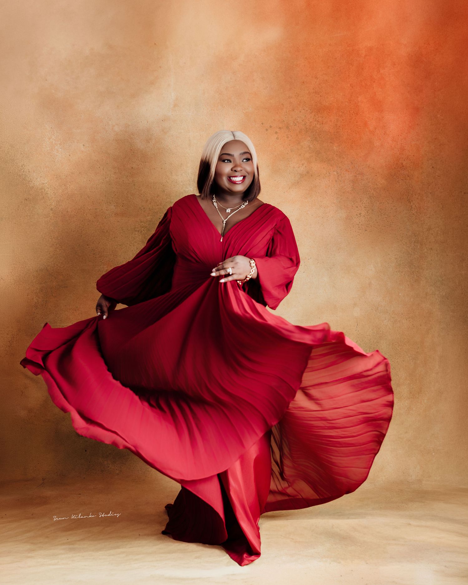 Woman in flowing red gown twirling gracefully in studio portrait with warm golden background.