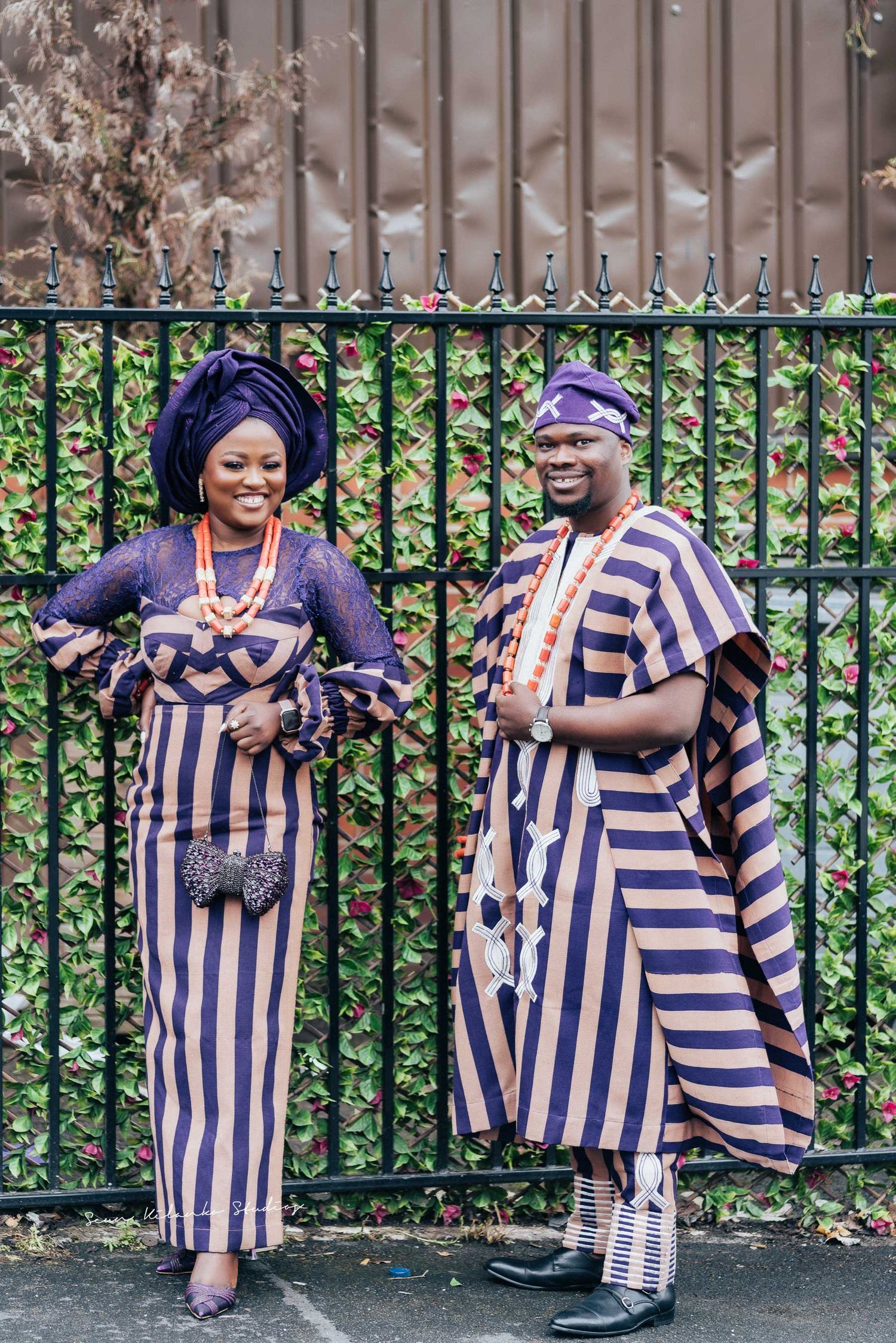 Two people in coordinating striped traditional African attire pose joyfully by iron fence.