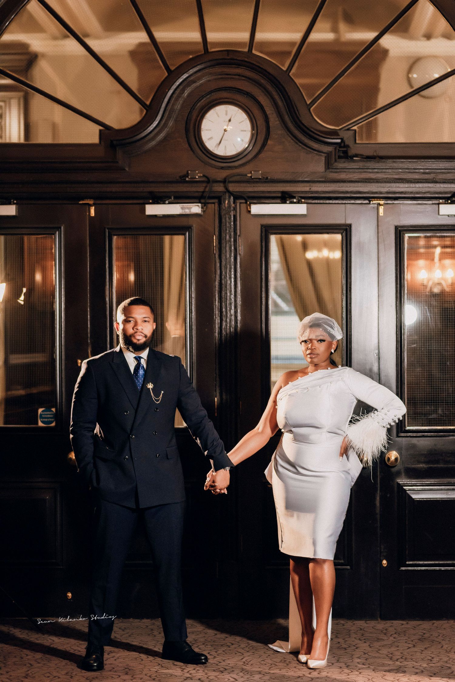 Elegant couple holding hands in formal attire before ornate dark wood doors with vintage clock at upscale venue.