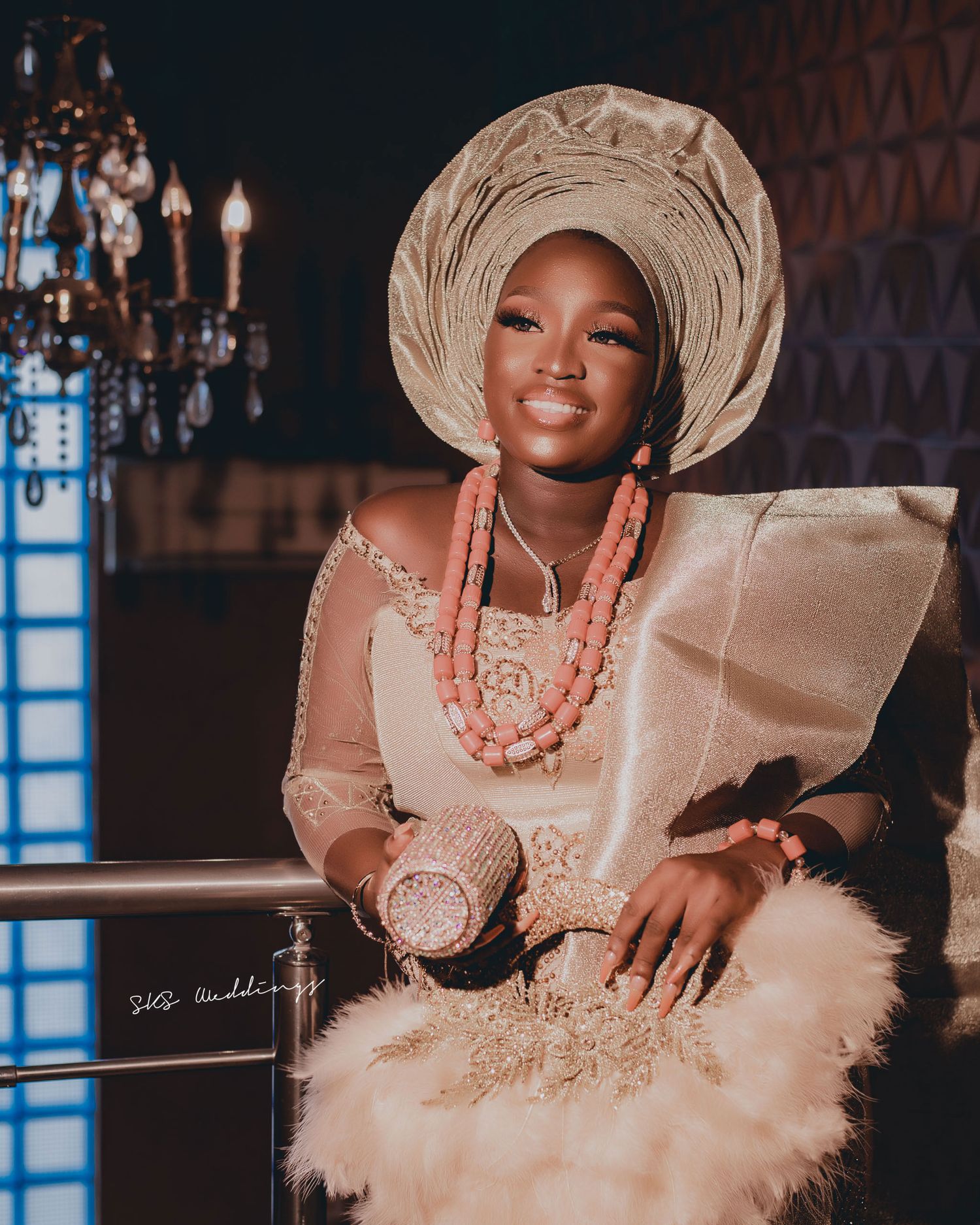 Traditional Nigerian bridal attire featuring cream gele headpiece, coral beads, and champagne dress in elegant indoor setting.