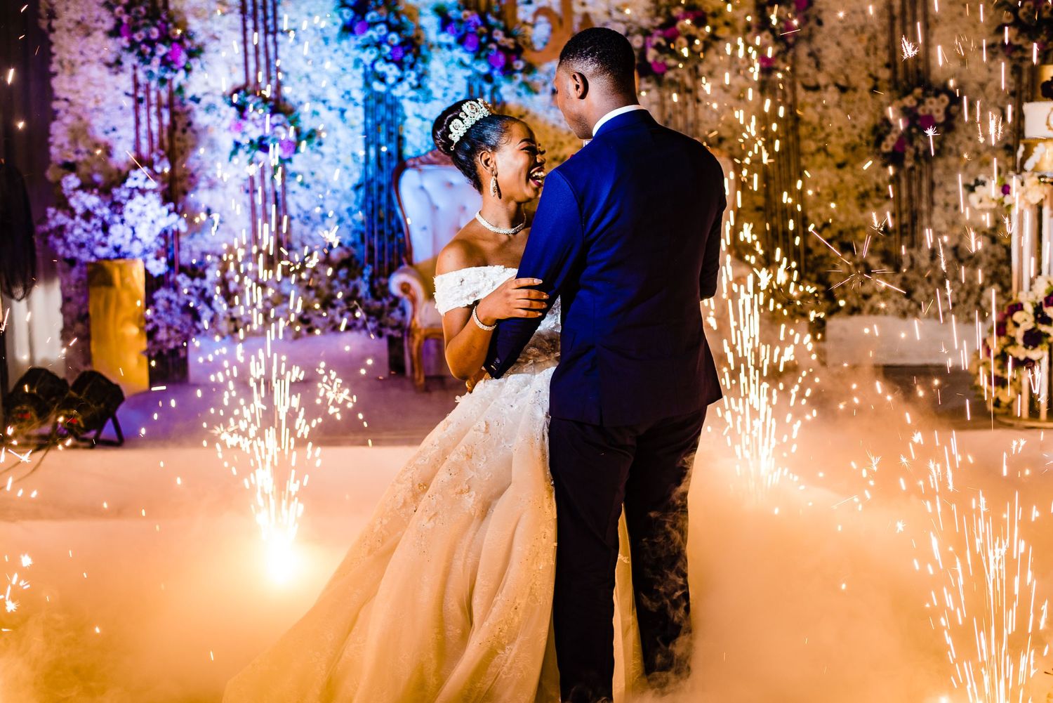 Couple shares their first dance at wedding reception with stunning sparkler fountains and colorful lighting effects in background.