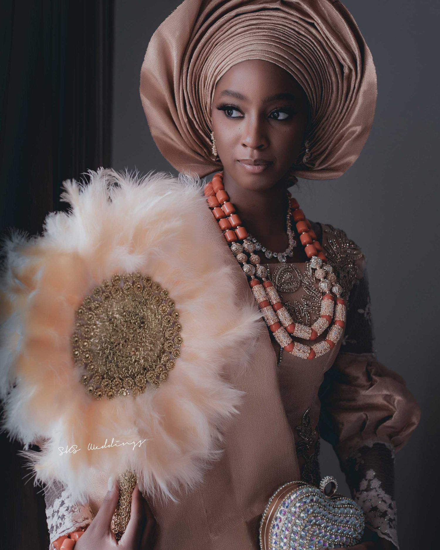Traditional African fashion portrait featuring a beige gele headpiece, coral beaded necklaces and a decorative white feathered fan.