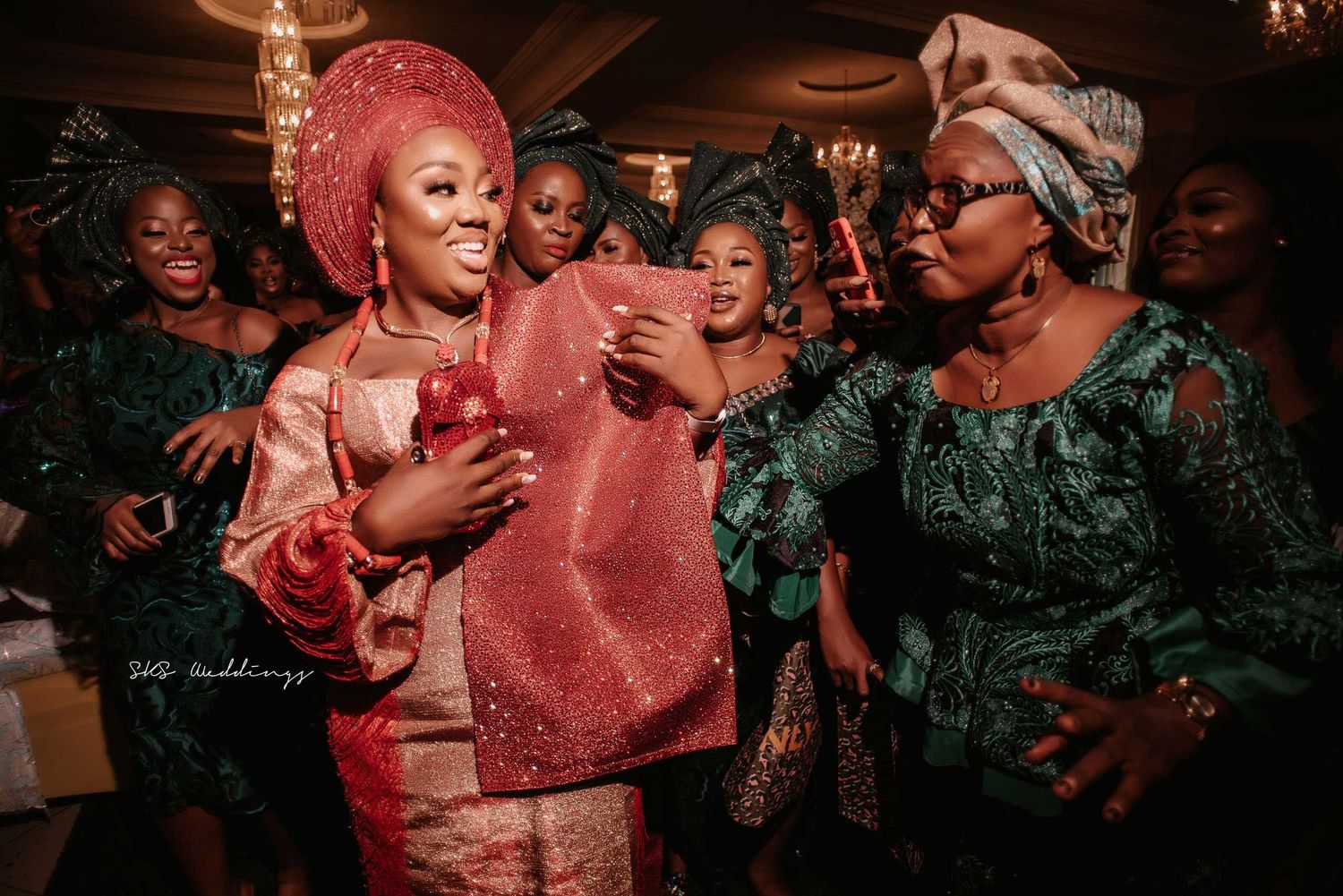 People celebrating at a lively party wearing traditional Nigerian attire in green and pink gele headwraps and dresses.