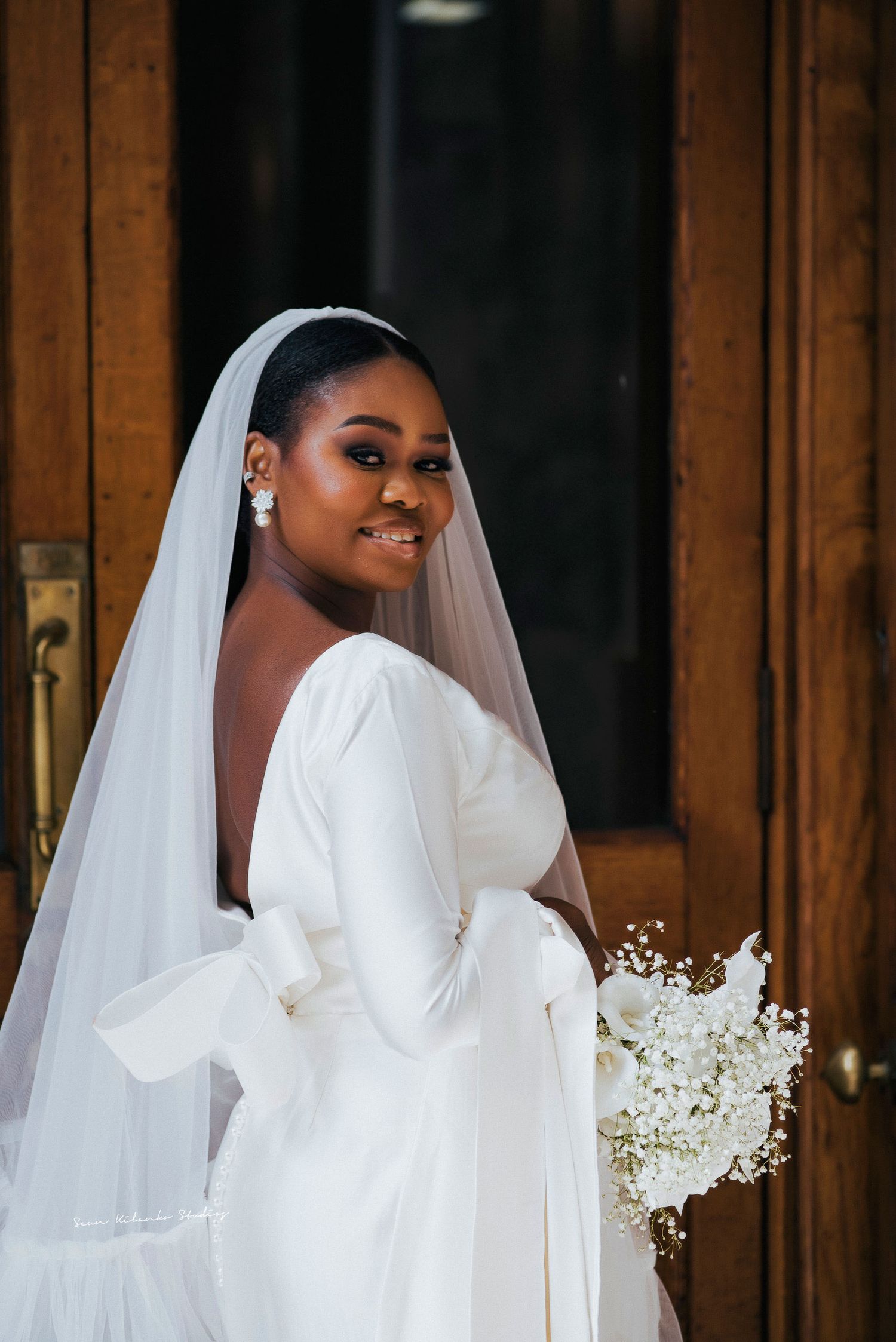 Elegant bride in long-sleeved wedding dress holding white bouquet against wooden door.