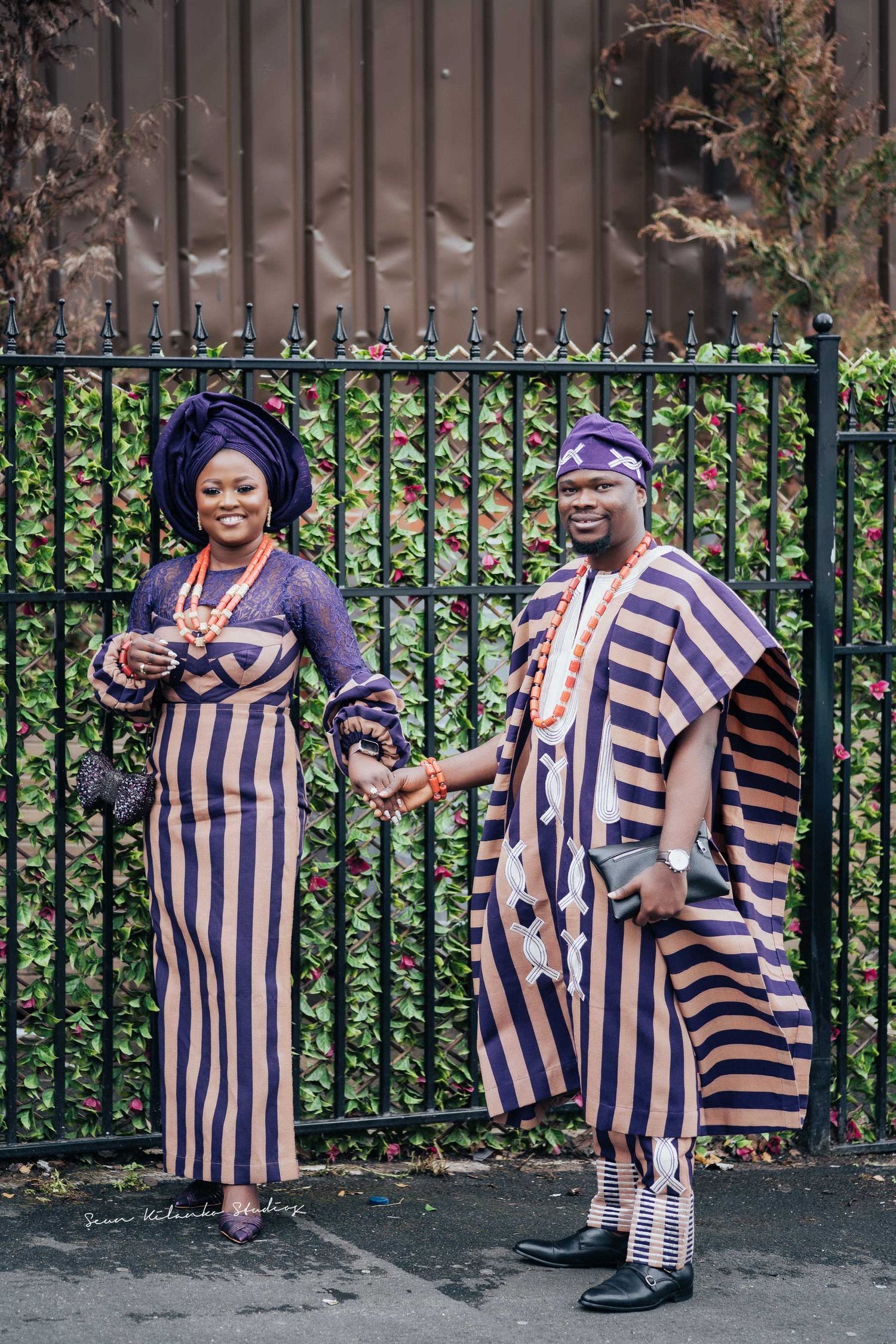 Stylish duo in complementary striped African outfits pose playfully against garden fence.