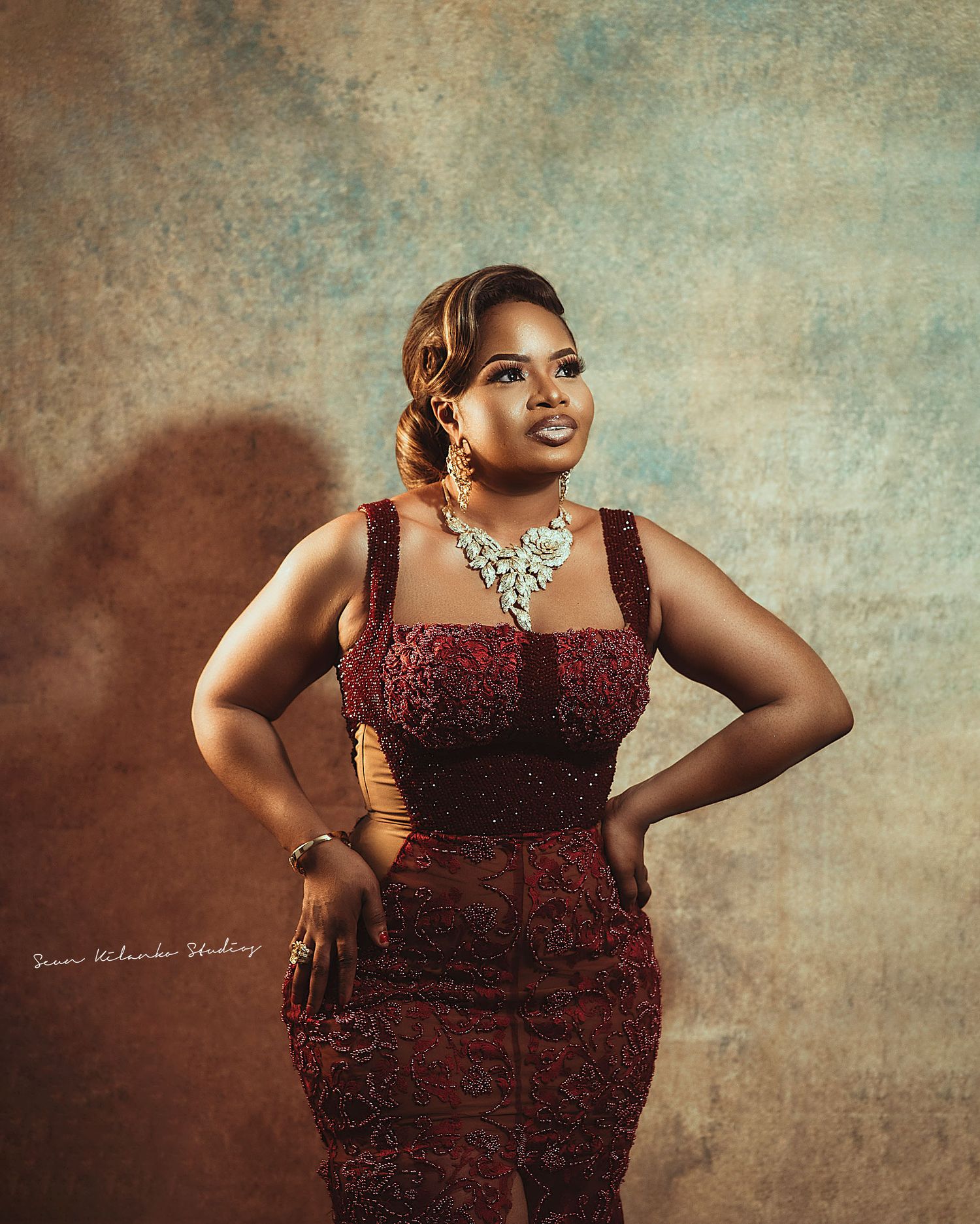 A glamorous portrait featuring a burgundy lace gown and statement crystal necklace against a textured backdrop.