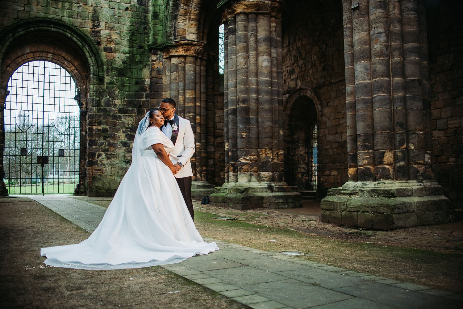Bride and groom embracing in romantic wedding photo among ancient stone ruins with arched columns.