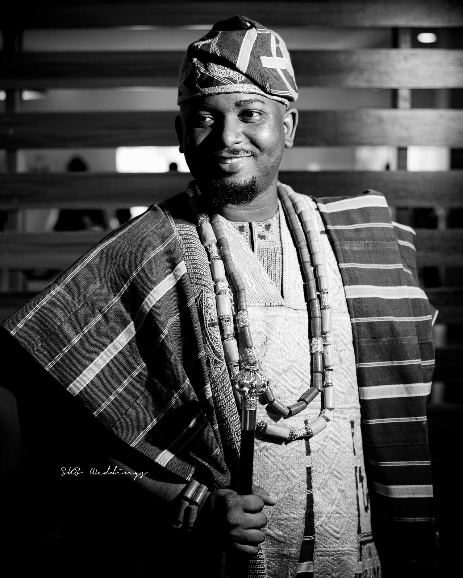 A black and white portrait of someone wearing traditional Nigerian Yoruba attire including an agbada and beaded necklaces.