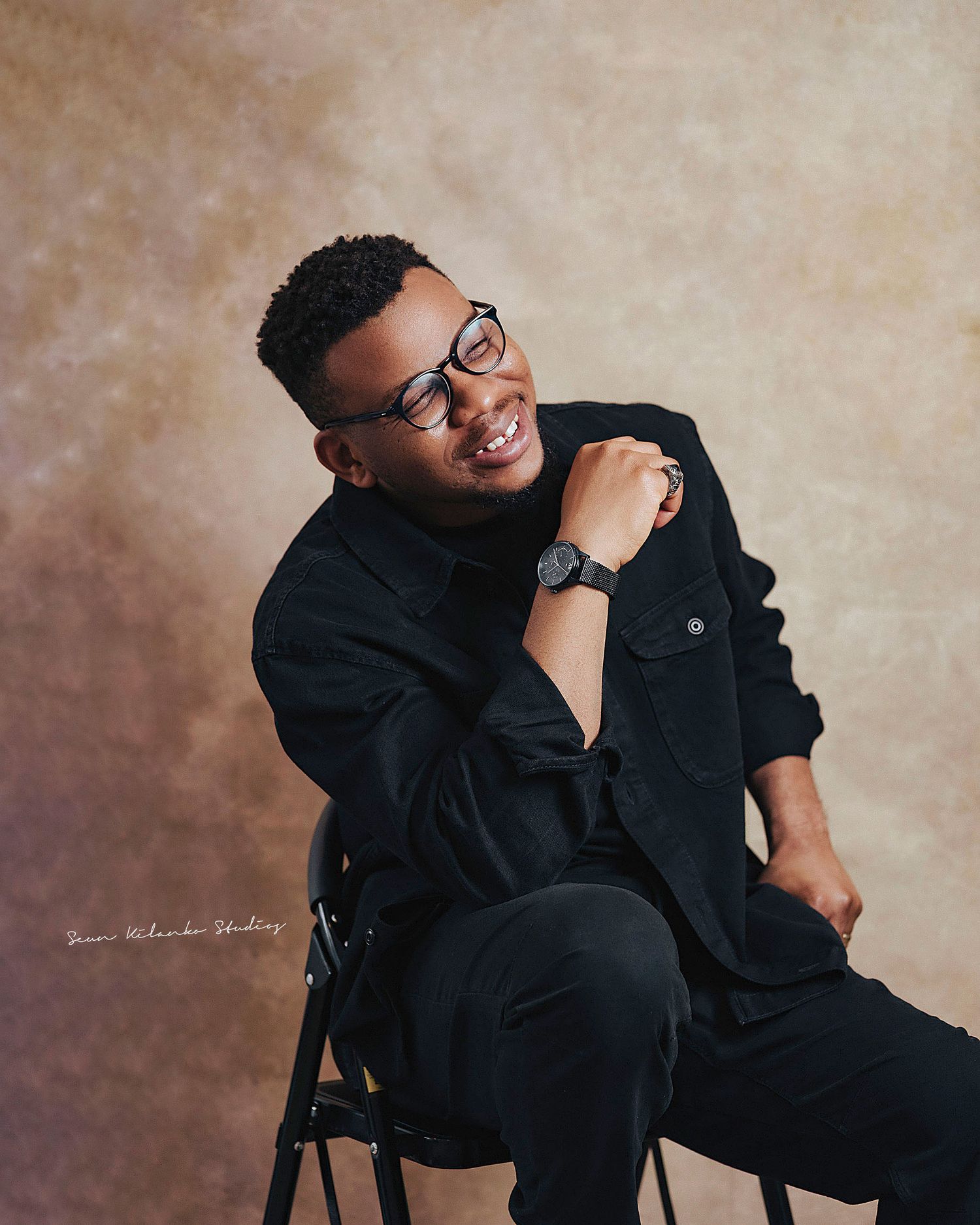 Joyful studio portrait of a person in all black attire seated on chair against neutral background.