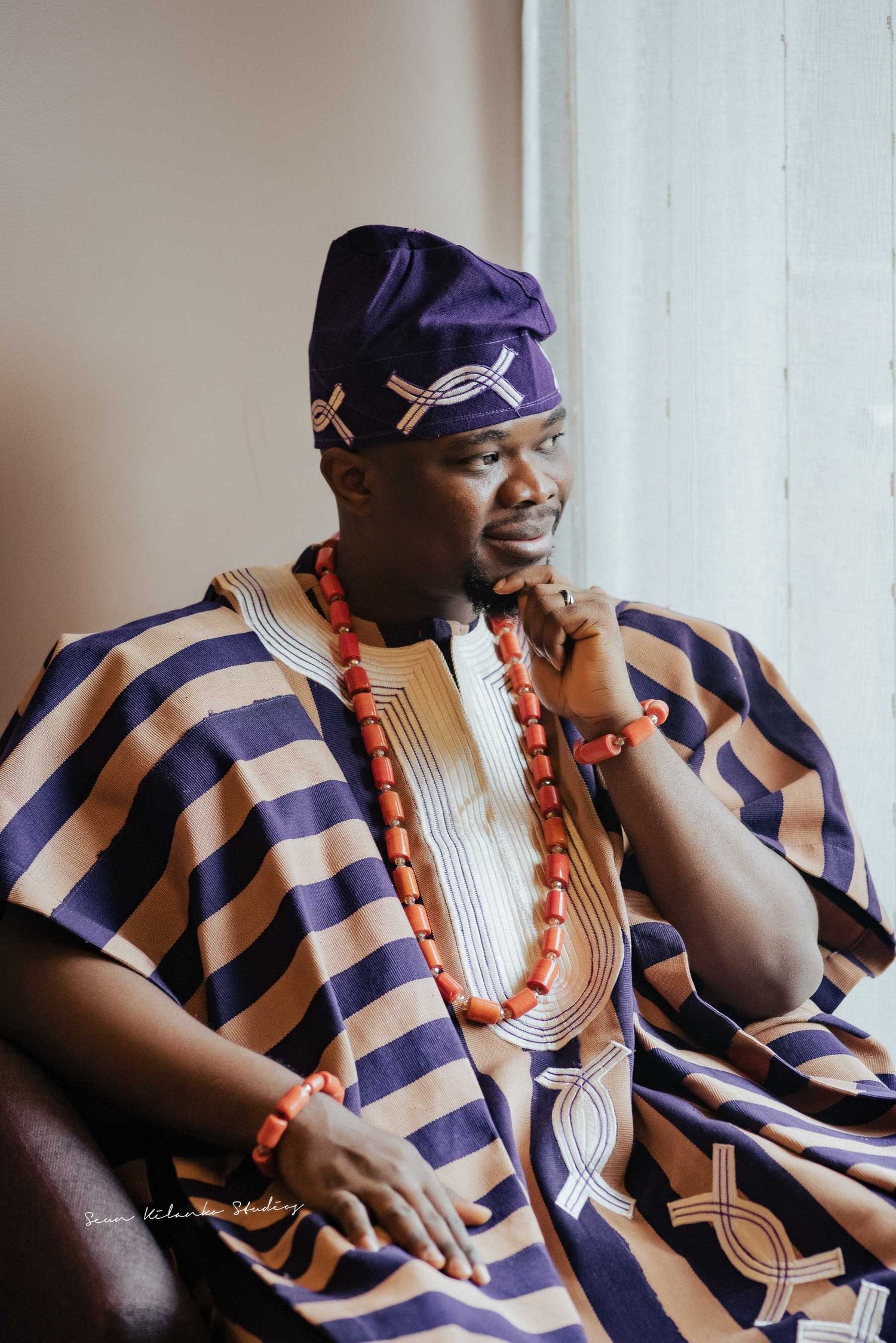 Portrait of someone wearing traditional Nigerian striped agbada and cap while sitting thoughtfully.