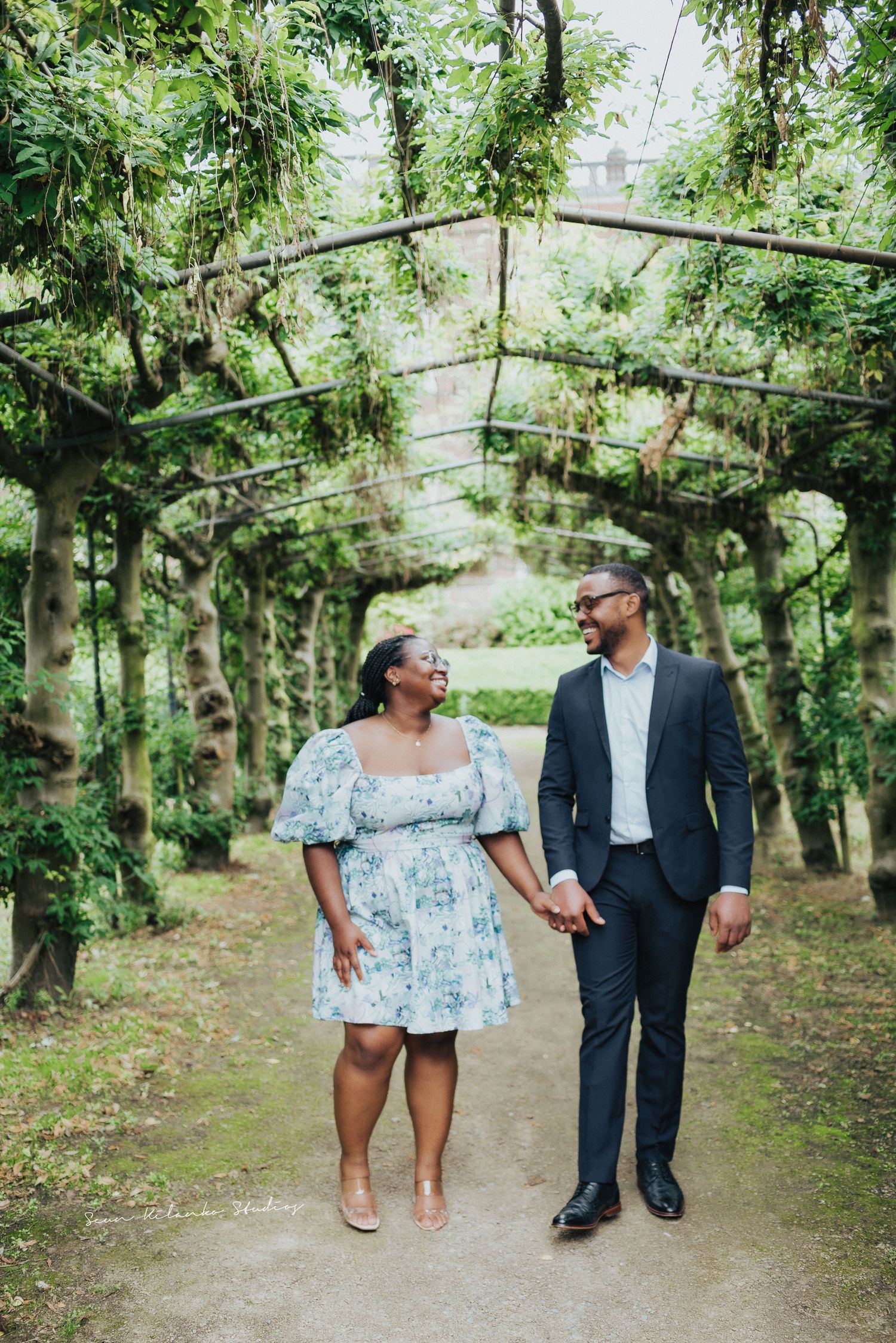 Couple holds hands while walking through lush garden archway.