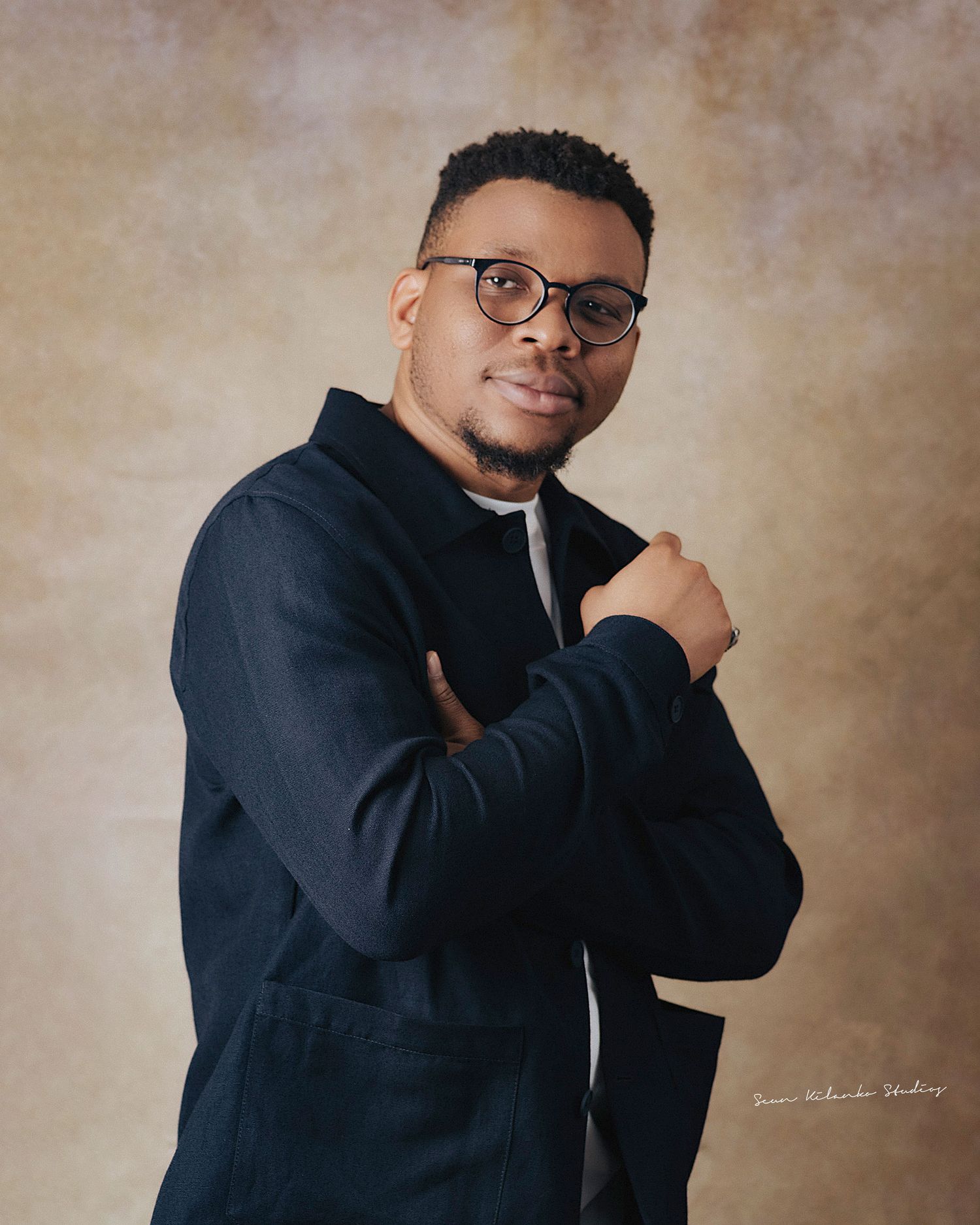 Studio portrait showing someone in a dark jacket and glasses with a warm smile.