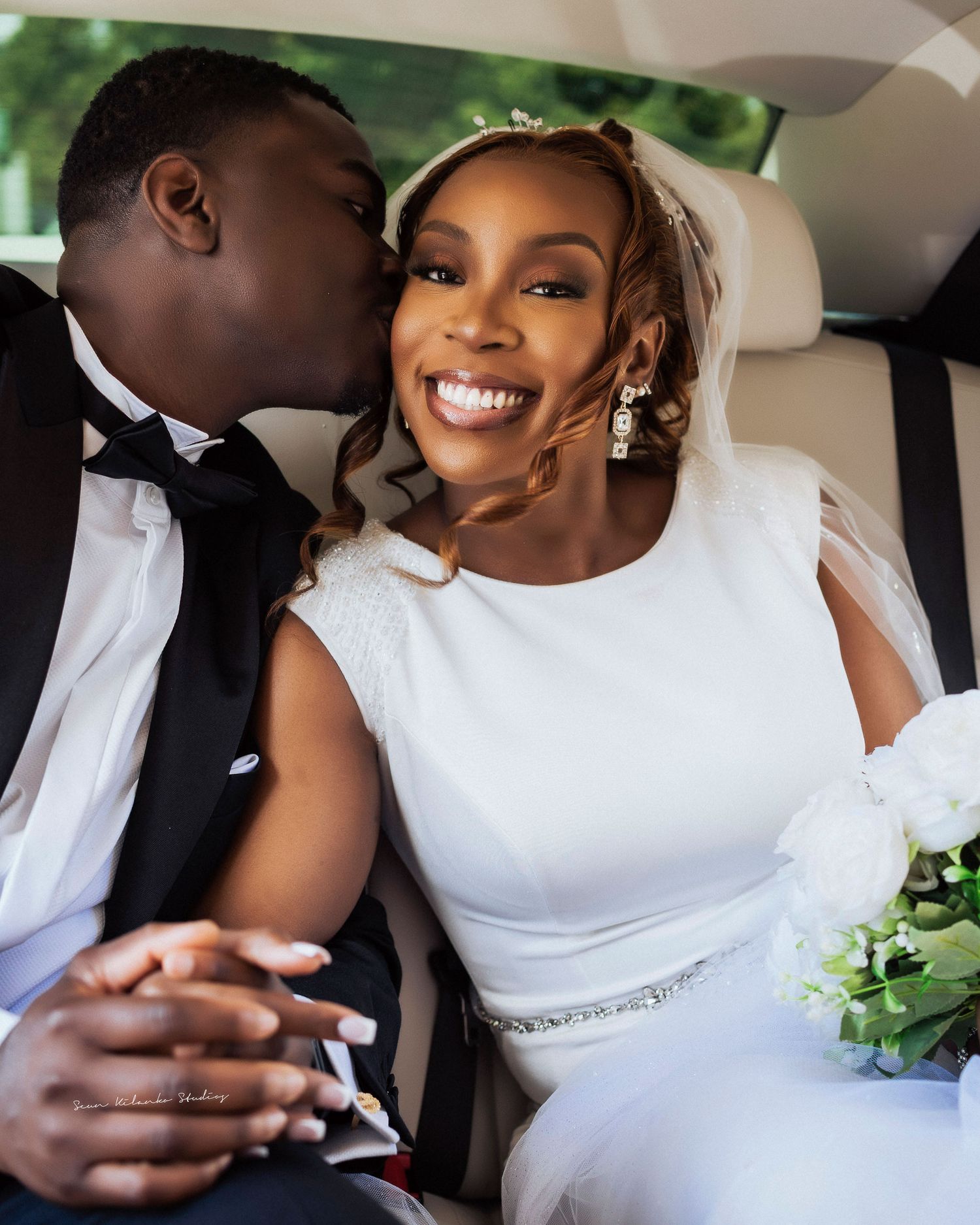 A joyful bride in a white gown smiles as her groom in a tuxedo kisses her cheek inside a wedding car.
