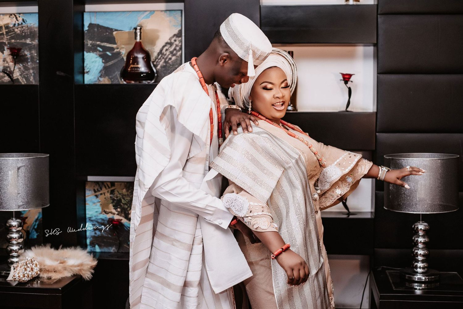 A couple in matching white traditional African attire pose elegantly against a modern interior backdrop.