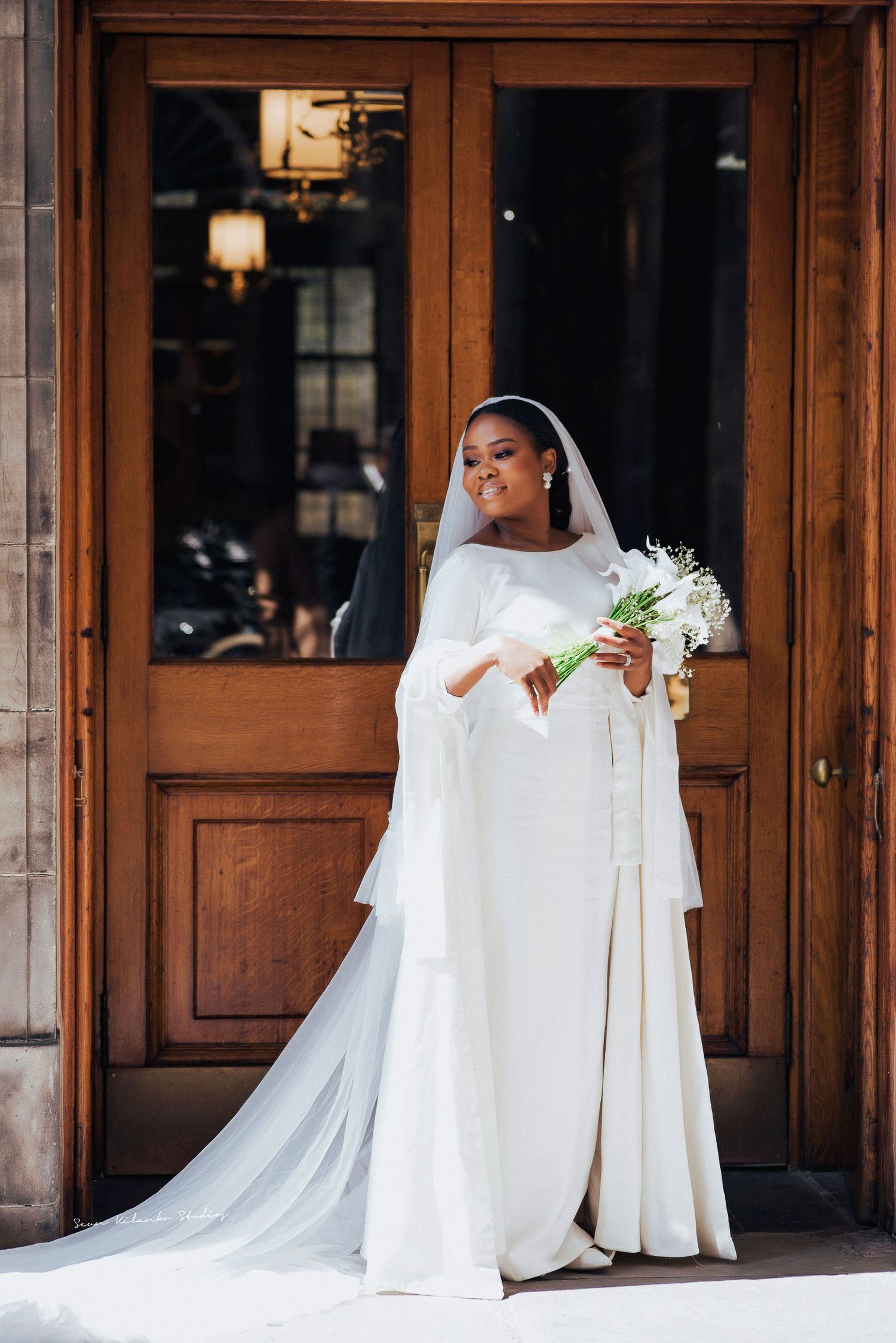 Elegant full-length bridal portrait with flowing dress and white bouquet against wooden door.