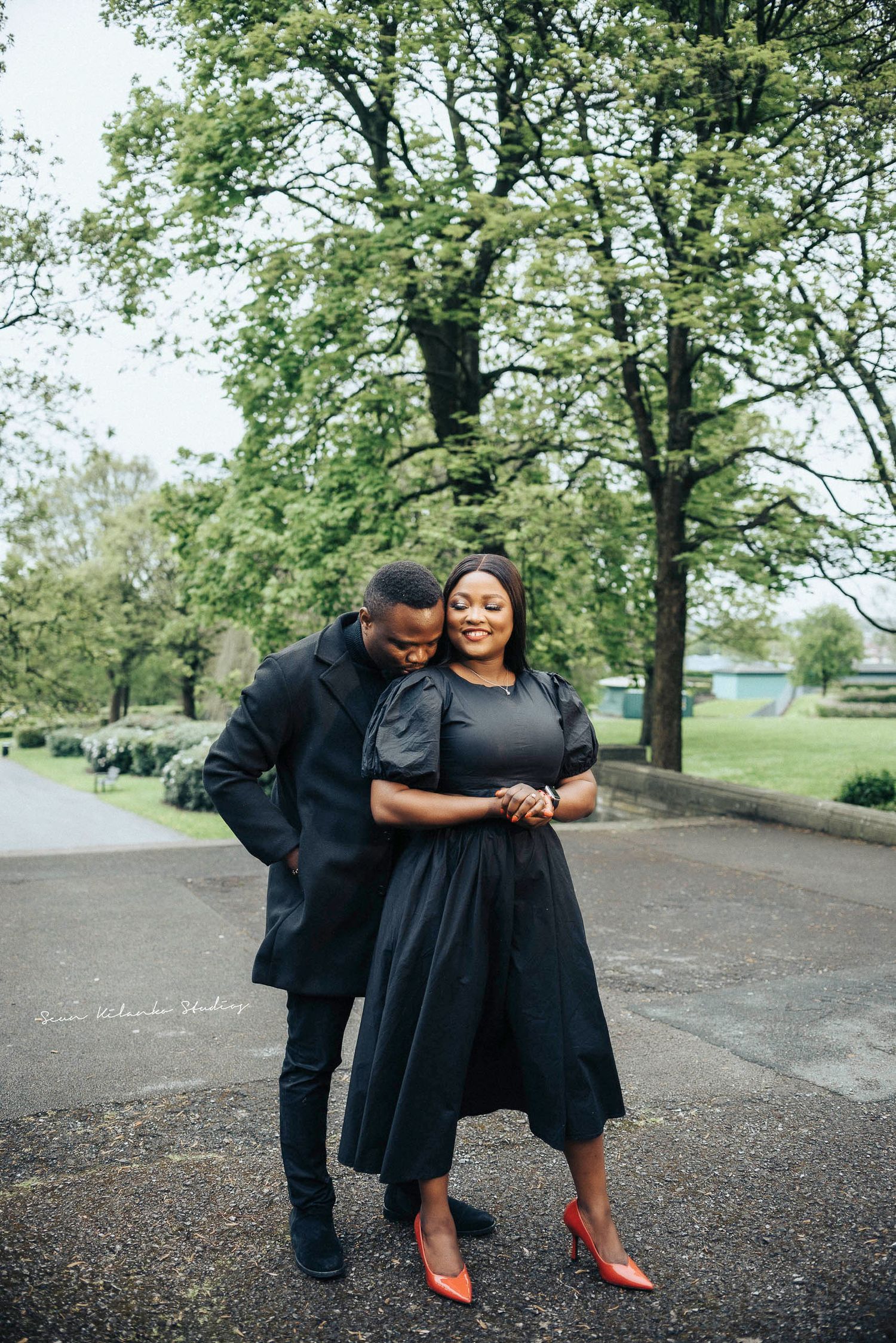 Couple embraces on path under spring trees wearing coordinated dark outfits.