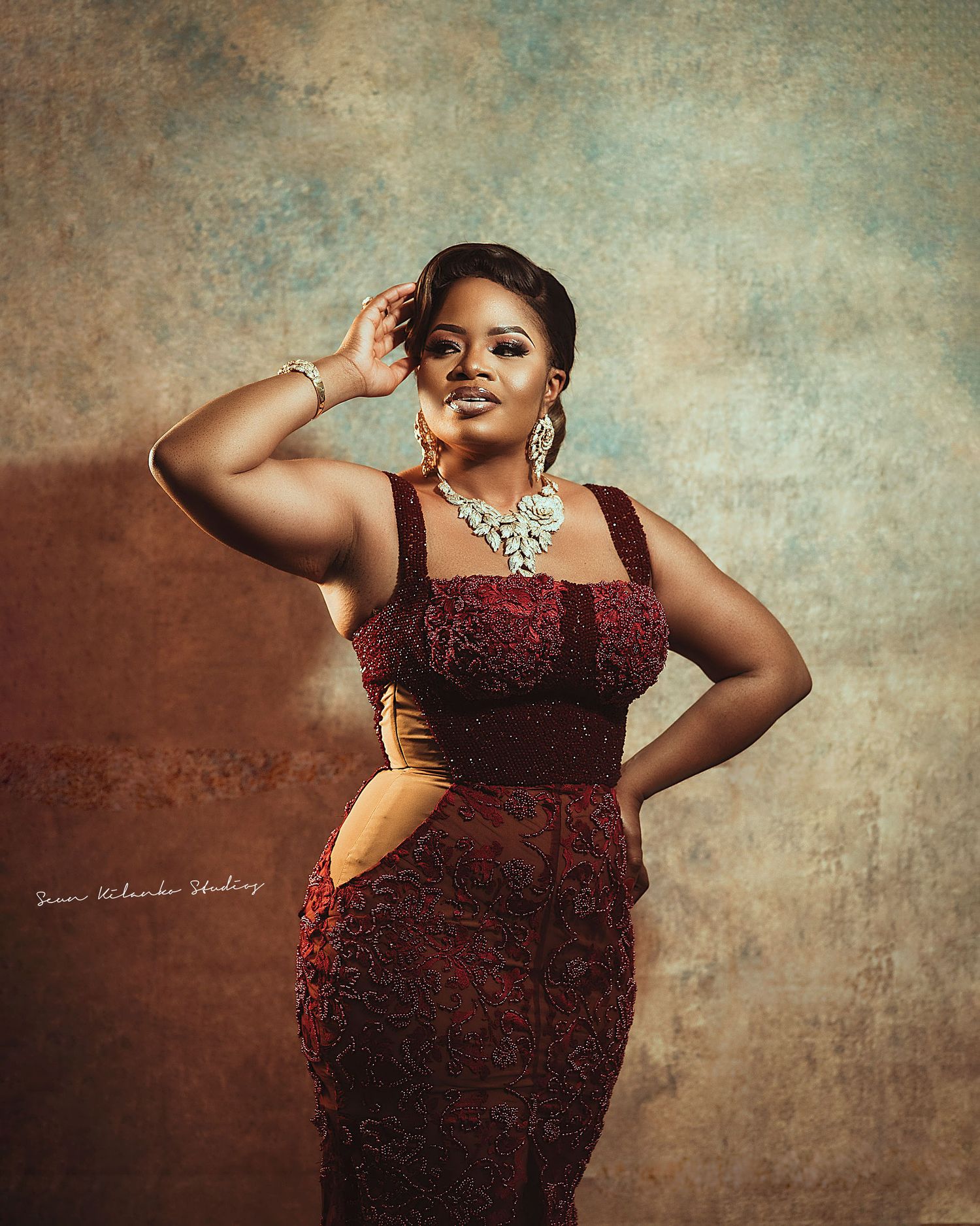Glamorous portrait pose in a burgundy lace dress with statement jewelry and styled hair.